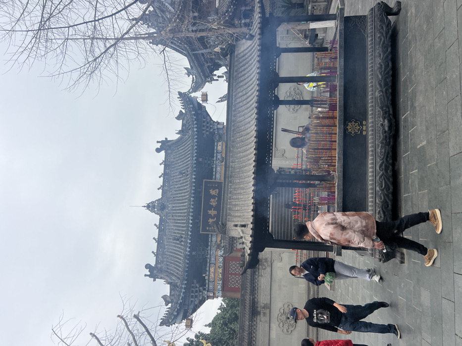 The image depicts an outdoor scene within Ciyun Temple (慈云寺), located in Shapingba District, Chongqing, China.
In the background, prominent traditional Chinese temple structures are visible, characterized by dark tiled roofs with ornate ridge decorations and dark eaves. The central building features a dark plaque with gold Chinese characters, "慈雲寶航" (Cí Yún Bǎo Háng). Below these buildings are light grey stone walls. The sky above is uniformly overcast. Bare deciduous tree branches are present on the left and right sides of the frame, suggesting a leafless season.
In the mid-ground, a large, black, rectangular incense burner (censer) stands under an open-sided pavilion with a dark tiled roof supported by four pillars. Numerous multi-colored incense sticks burn upright within the censer, producing a substantial amount of smoke that rises into the pavilion. The censer features intricate relief carvings along its base and a golden emblem on its front. The ground in the foreground and mid-ground is paved with light grey tiles.
Several individuals are present. In the foreground, a male wearing a light brown puffer jacket, dark trousers, and tan shoes walks from right to left, holding a clear water bottle. To his left and slightly behind, another male wearing a dark baseball cap, a dark long-sleeved shirt with white text, and blue jeans walks towards the left, accompanied by a partially visible child in a red jacket. Alongside them, a female in a dark hoodie and light grey pants walks left, carrying a green bag. Behind the censer, inside the pavilion, at least two females are visible: one is actively tending to the burning incense, and another is partially visible to the right, possibly wearing a yellow vest. Additional indistinct figures are present further in the background near the temple steps. The overall activity centers around the burning incense and pedestrian movement through the temple courtyard.