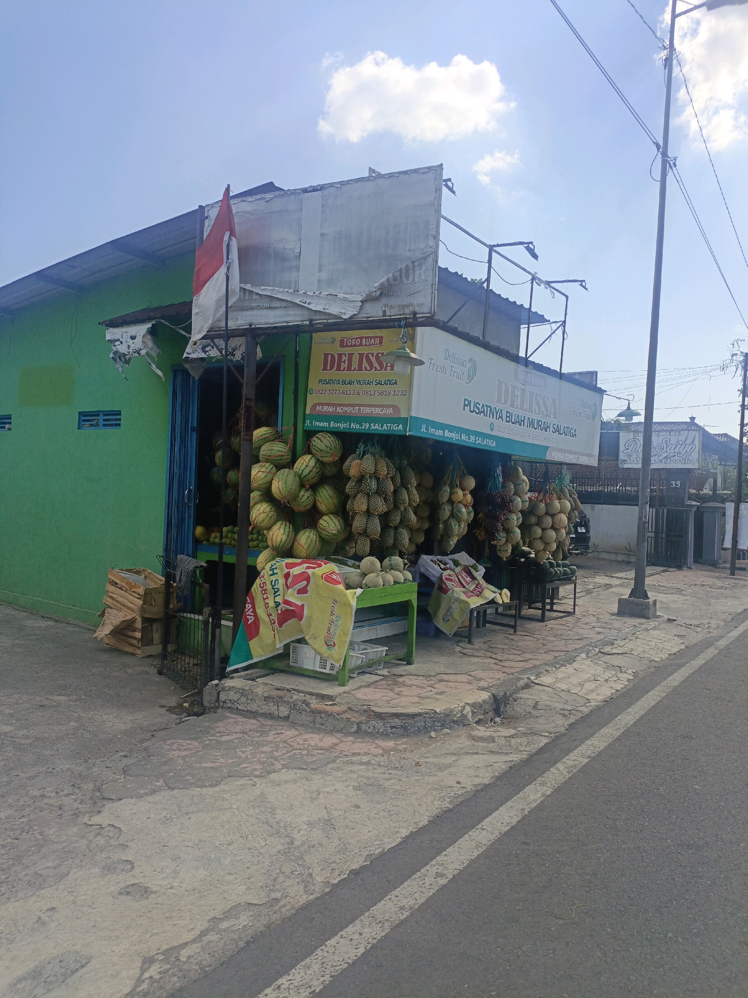 The image shows a fruit stall called "Delissa Fresh Fruit" located at Jl. Imam Bonjol No. 39 Salatiga, Indonesia. The stall is situated on a sidewalk next to a road. The stall is selling various fruits, including watermelons, pineapples, and durians, which are displayed on tables and hanging from the ceiling. A green building with blue window shutters is visible behind the stall. The Indonesian flag is flying from a pole in front of the stall. The sky is blue with scattered clouds. The road is paved and has a white line marking.