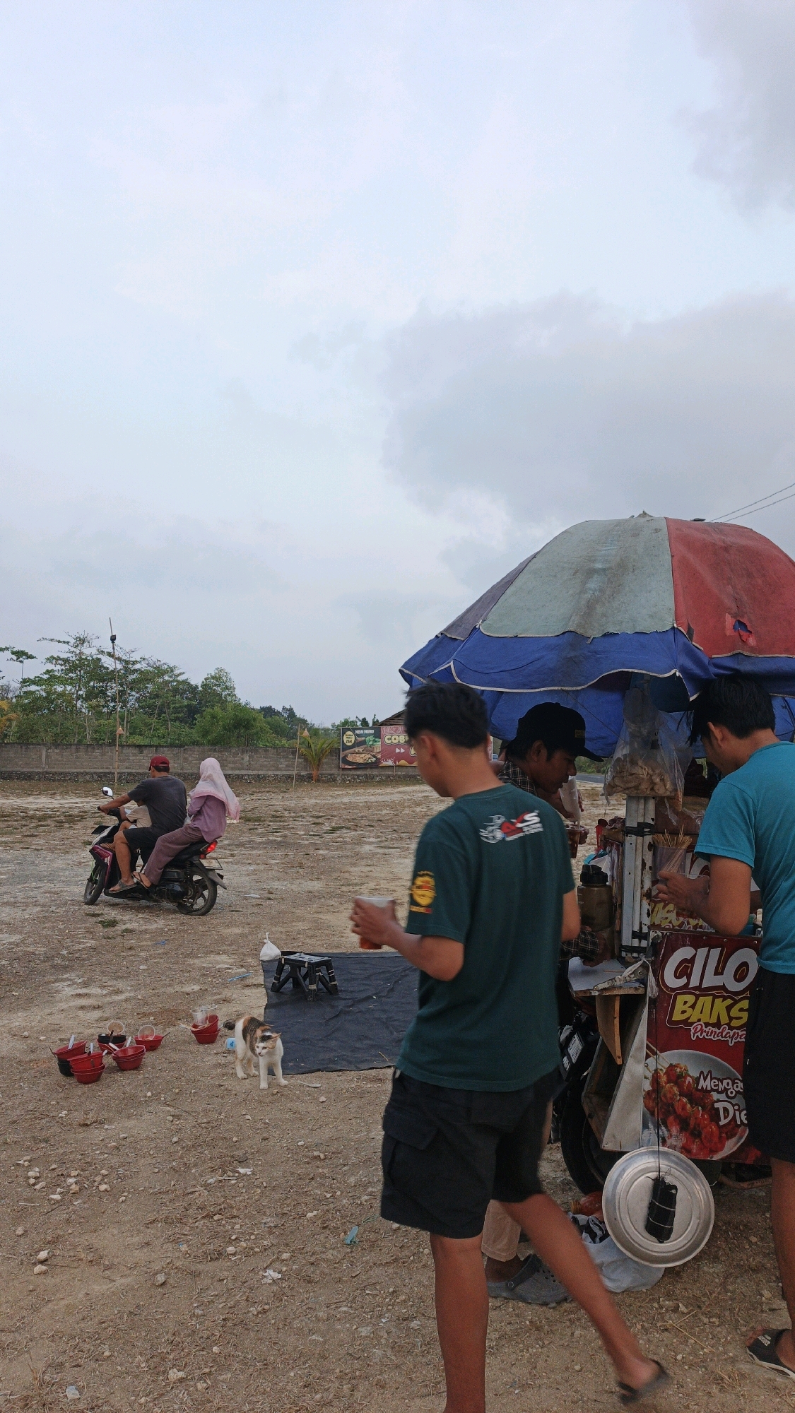 In Pangandaran, Indonesia, a street food cart is set up under a large, multi-colored umbrella. The cart displays a sign advertising "CILO BAKSO Prinsipa" with an image of food. Three individuals are interacting around the cart; one person in a green t-shirt and black shorts holds a drink and faces away from the camera, another person in a blue t-shirt stands at the side of the cart, and a third person wearing a black cap is behind the cart, seemingly serving customers. In the background, a motorcycle with two passengers, one wearing a red cap and the other a pink hijab, rides away from the cart. Several red buckets are lined up on the sandy ground, and a calico cat stands near them. The environment appears to be an open, dusty area with some vegetation and a brick wall in the distance. The sky is overcast with clouds.