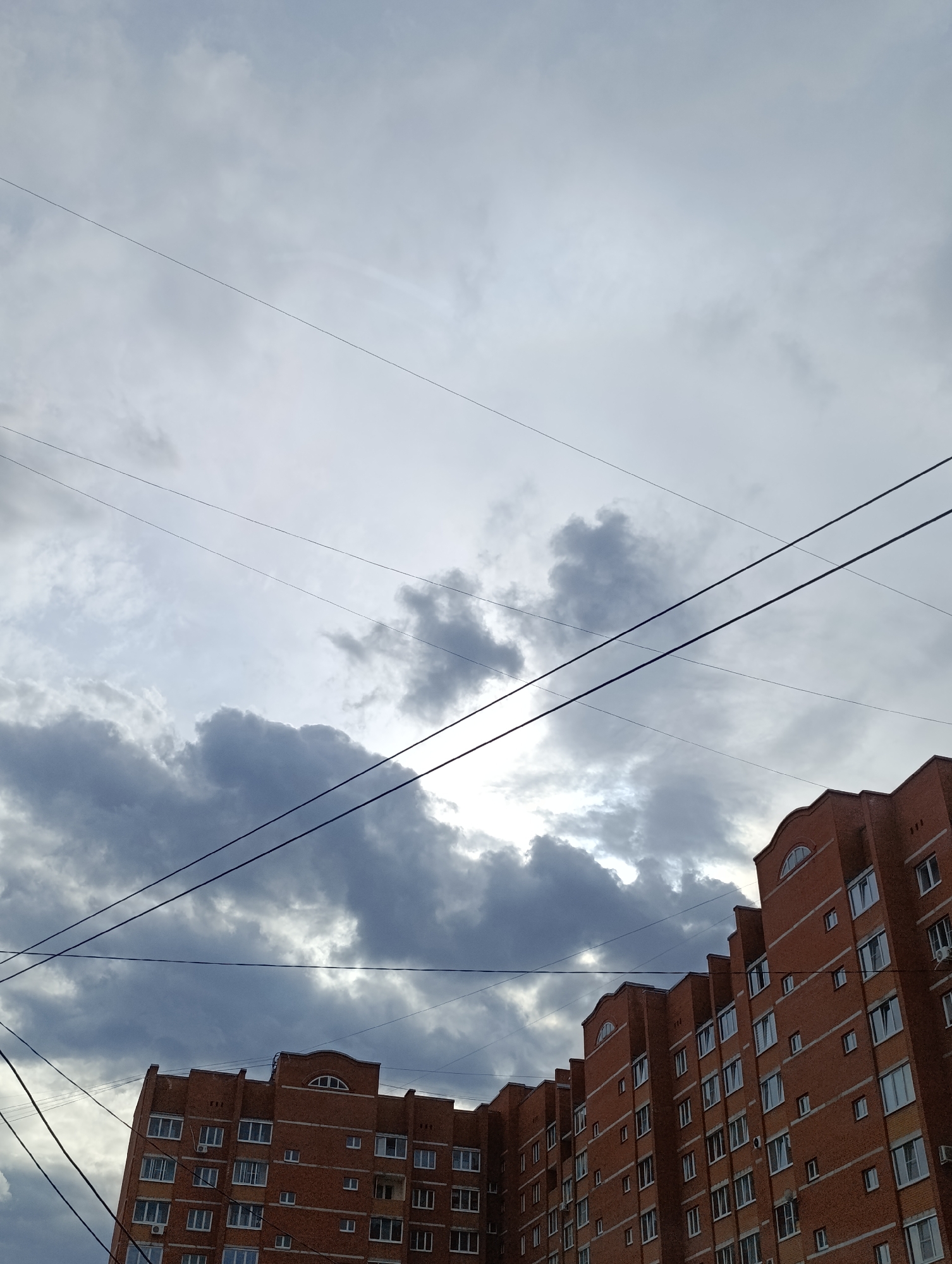 This vertical shot captures an urban scene dominated by a large, red brick apartment building under a heavily overcast sky. The multi-story building, likely residential, features numerous rectangular windows with white frames, many of which have individual air conditioning units. Its architecture is distinctive, with a stepped facade and arched windows on the upper sections, characteristic of residential buildings in places like Chaikovsky, Russia.
The sky occupies a significant portion of the frame, filled with a mix of light and dark grey clouds. A bright patch of light is visible behind a darker cluster of clouds, suggesting the sun is attempting to break through or is behind the cloud cover, indicating daytime. Multiple dark utility wires crisscross the upper and middle portions of the image, running diagonally across the sky and in front of the building, adding a layer of visual texture to the scene.
No people or explicit activities are visible, presenting a static snapshot of urban life. The weather appears cloudy, possibly hinting at an impending rain or just a generally overcast day. The overall impression is one of everyday urban scenery under a dramatic, cloudy sky.