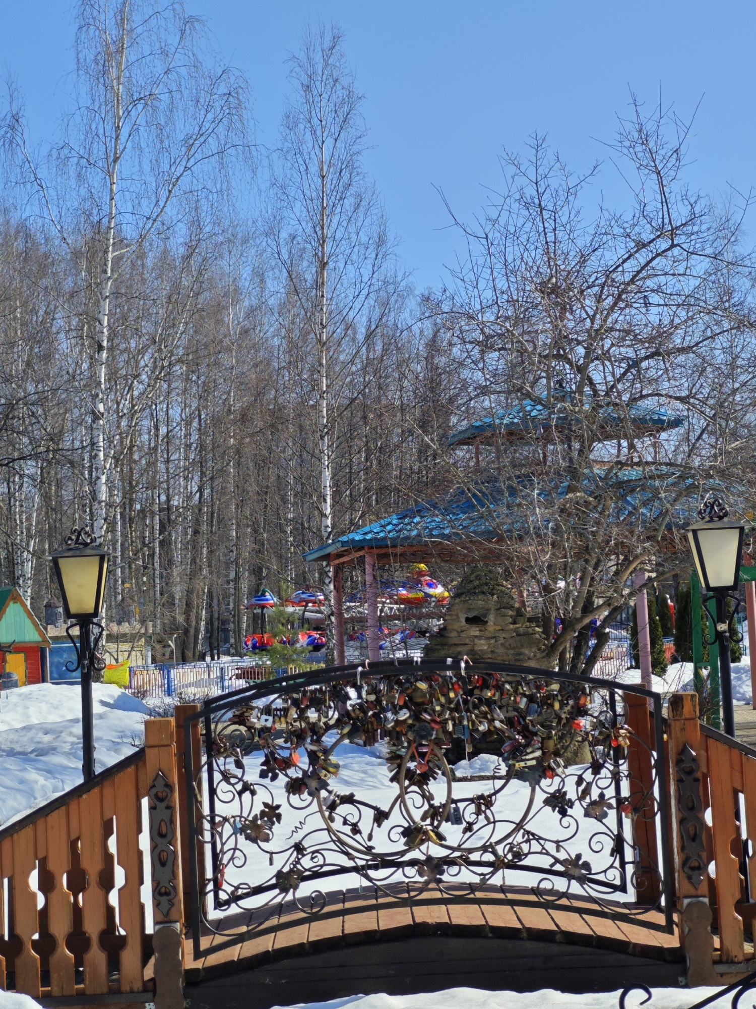 The image captures a scene in the city of Chaikovsky, Russia, showcasing a small ornate pedestrian bridge. The bridge features a dark metal railing decorated with a floral pattern and covered with numerous padlocks, suggesting a tradition of couples locking padlocks as a symbol of their love. The bridge itself has wooden sides with cut-out designs and leads over a snow-covered area. In the background, a park-like setting is visible with bare trees, a three-tiered gazebo with a blue roof, and a colorful children's ride. Two vintage-style lampposts flank the bridge. The sky is clear and blue. The presence of snow indicates it is winter or early spring.