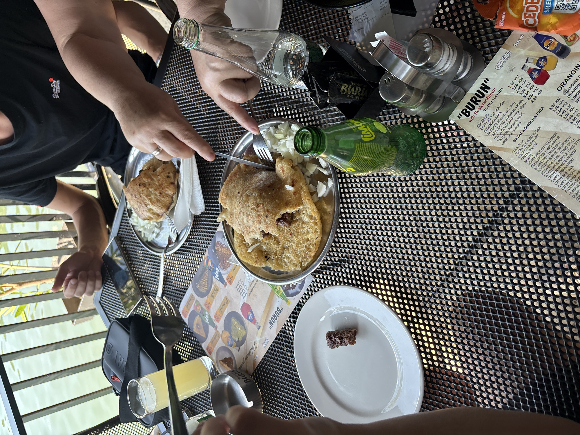 This is a close-up, eye-level outdoor shot of a table at a restaurant with a waterfront view, likely in Divic, Bosnia and Herzegovina. A group of people are engaged in eating and drinking. In the foreground, a white plate holds a single, small piece of dark brown meat. To the right, a silver metal salt and pepper shaker set sits on the table. A green glass bottle of "Limona" soda is positioned next to it. A black menu with "BURUN" and "Wifi. Restoran Burun" printed on it is also visible on the table. In the middle ground, a person's hands are visible holding a fork and knife, cutting into a dish of flatbread filled with meat and topped with chopped onions, served on a silver platter. Another silver platter with a similar dish is to the left, alongside a glass of light-yellow liquid and an ashtray. In the background, a person in a black t-shirt is seated, with their hands resting on the table. A black bag with a red zipper is placed on the table. The table surface is a black, perforated metal mesh. A metal railing and calm water are visible in the background, suggesting a waterside location.