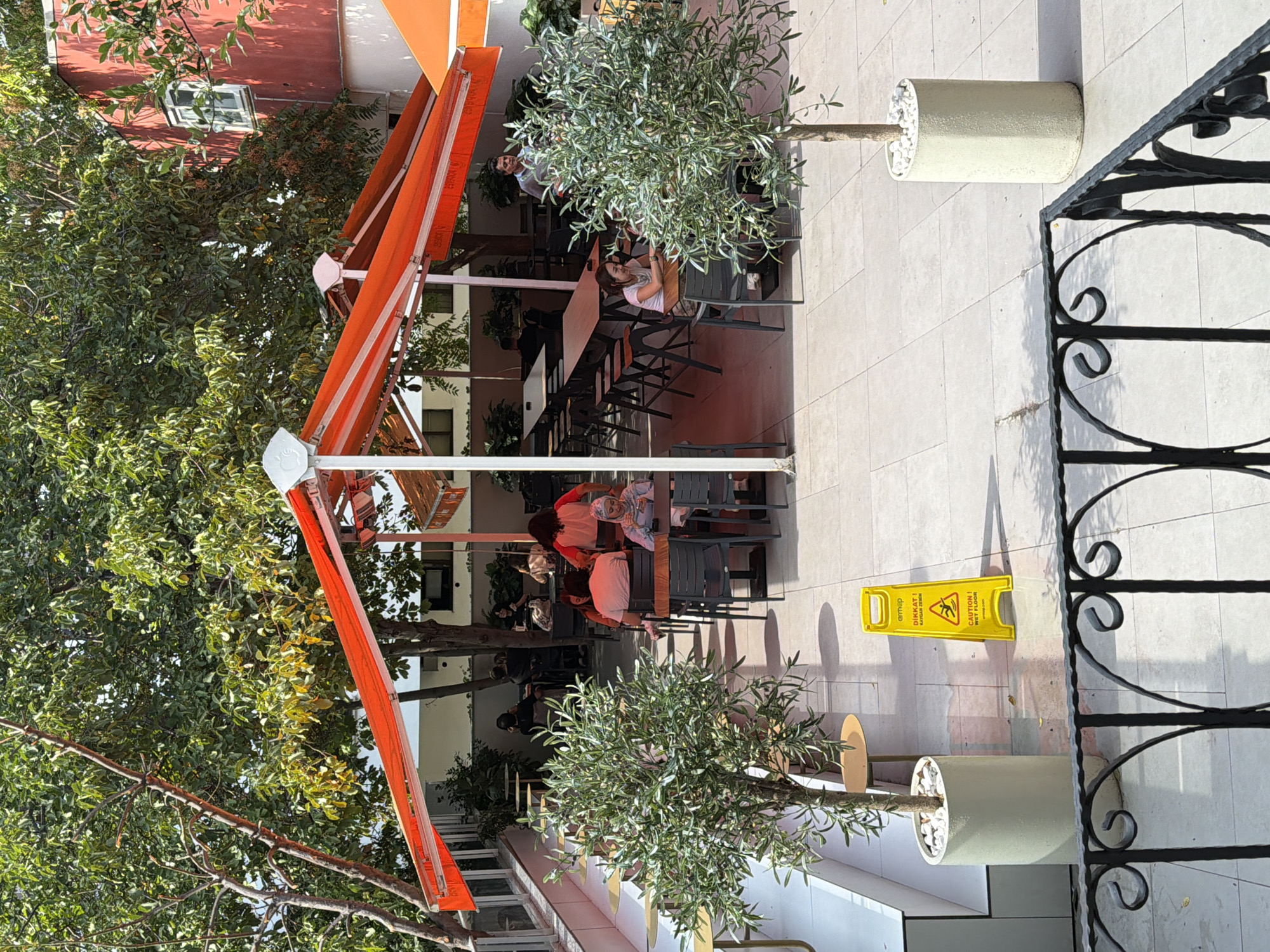 The image depicts an outdoor seating area of a cafe, likely in Kadıköy, Turkey, as indicated by the presence of a "DİKKAT! KAYGAN ZEMİN / CAUTION! WET FLOOR" sign in Turkish and English. The area is furnished with numerous tables and chairs, some covered by an orange and white striped awning. Several people are seated at tables, engaged in dining or conversation. Two potted olive trees flank the seating area. In the background, a red building with a window is visible. The ground is paved with light-colored tiles, and shadows suggest a sunny day. A black metal railing is in the foreground.
