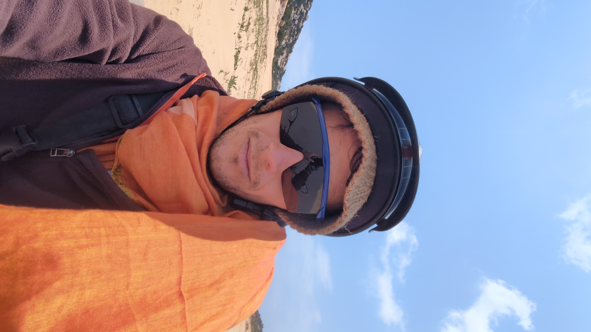 A male person is wearing sunglasses and a helmet, potentially participating in an outdoor activity such as paragliding. The background consists of a light blue sky with some clouds and a landscape with grassy terrain or dunes. The location is likely near Phuoc Dinh, Vietnam, based on the environment and the general appearance. The person is also wearing an orange scarf and layered clothing, including a brown fleece and a black strap.