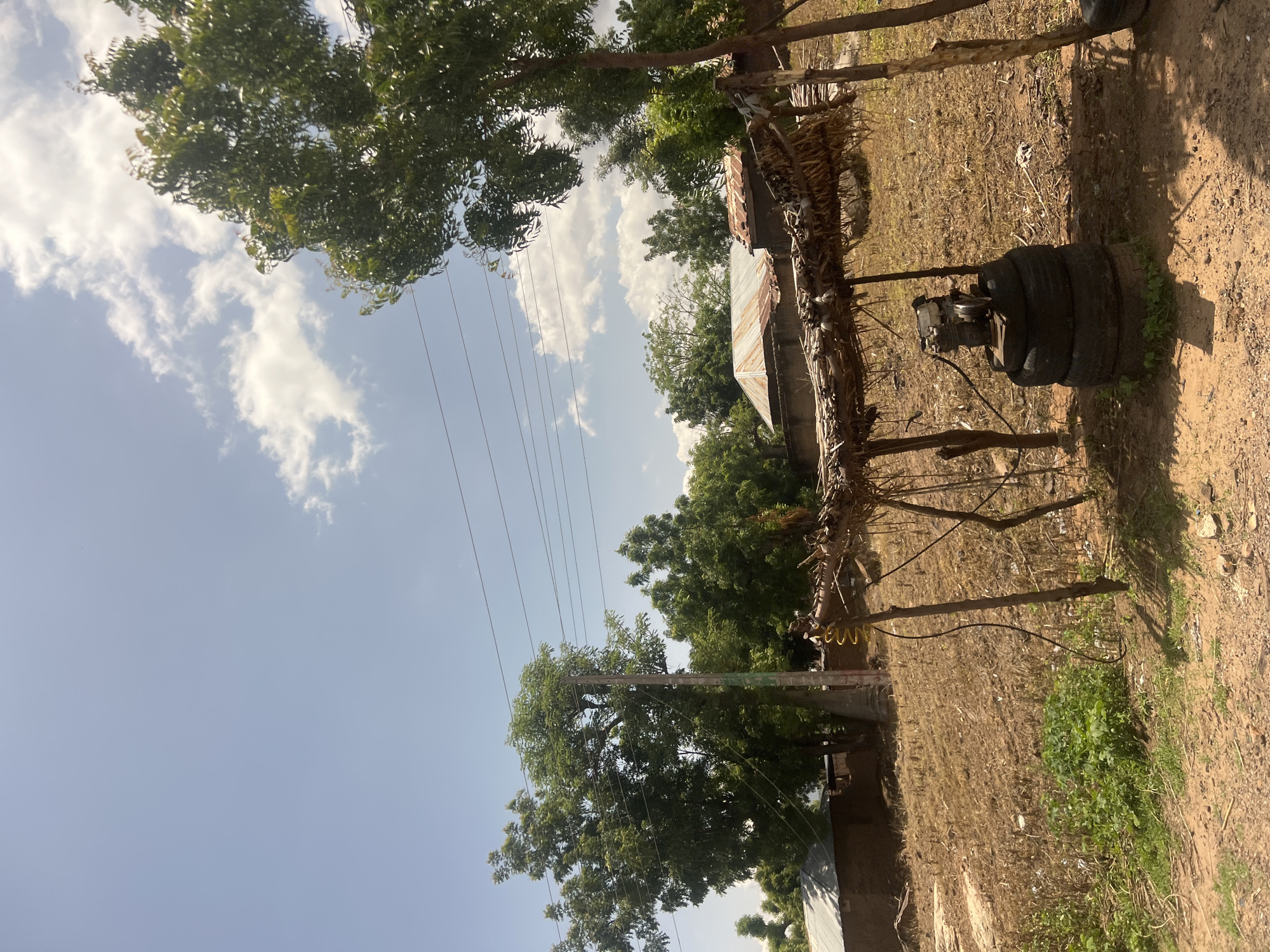 The image portrays an outdoor scene in Chum, Nigeria, under a bright, partly cloudy sky. No individuals are visible. In the right foreground, a rudimentary shelter is constructed from wooden poles supporting a roof of dried leaves or thatch. Beneath this shelter, a small engine, possibly a generator or pump, rests on a stack of three black rubber tires. A coiled yellow cable and a black cable are connected to the engine setup. The ground throughout the scene consists of dry, reddish-brown soil with sparse patches of green vegetation and dried grasses. In the midground, several single-story structures with mud walls and corrugated metal roofs are present. A tall utility pole, seemingly made of concrete, is located in the left-center, supporting multiple horizontal power lines. Numerous trees with dense green foliage are distributed across the midground and background, interspersed with the structures. The sky is bright blue with scattered white cumulus clouds.