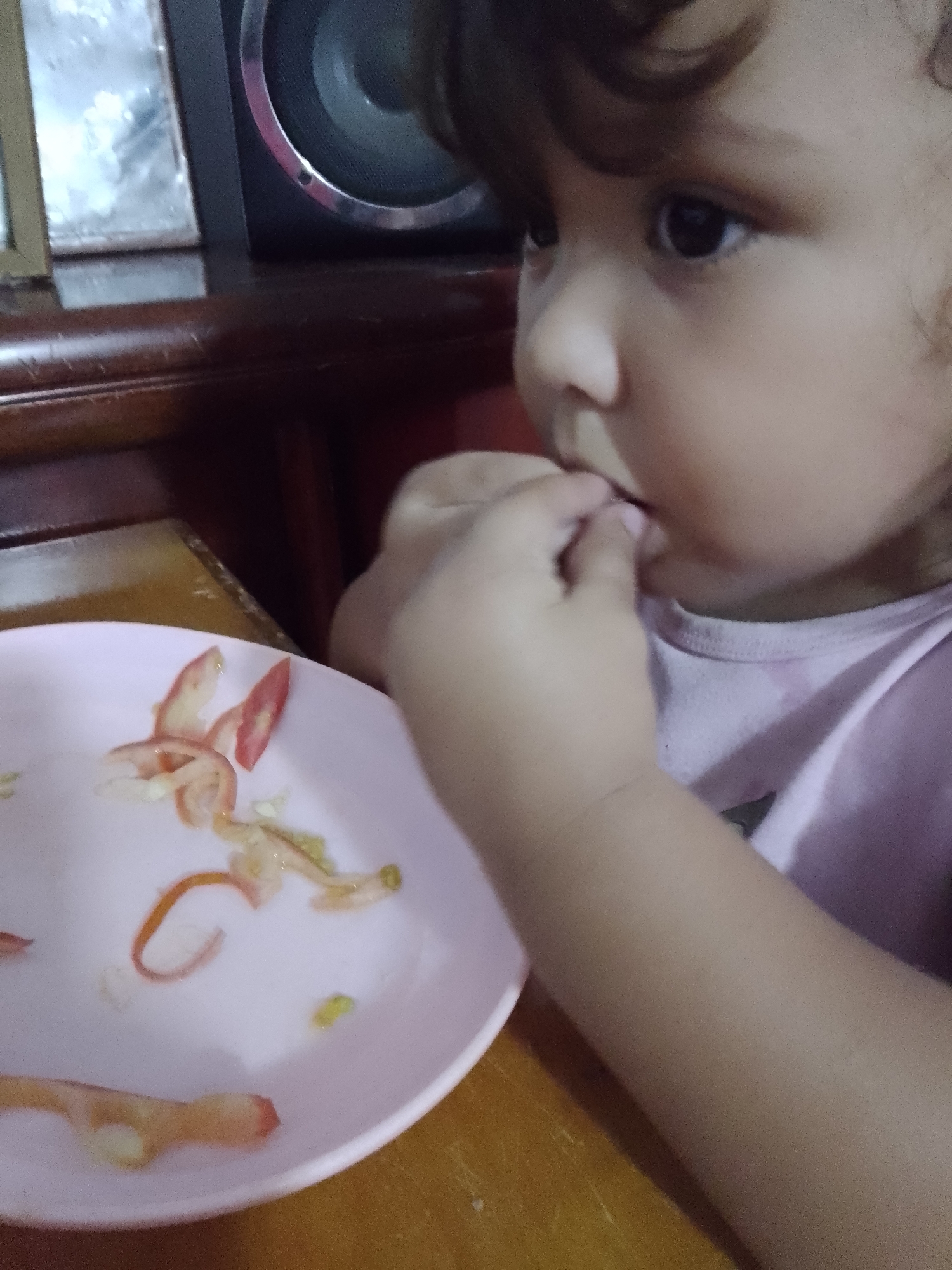 A young child is eating food from a light pink plate. The plate contains remnants of what appears to be tomato and possibly onion or cucumber. The child is holding food to their mouth with their right hand. The child is wearing a light pink shirt. A dark-colored speaker or electronic device sits in the background. The setting seems to be indoors, likely at a table. There is no information to verify the location of this image in Caracas, Venezuela.