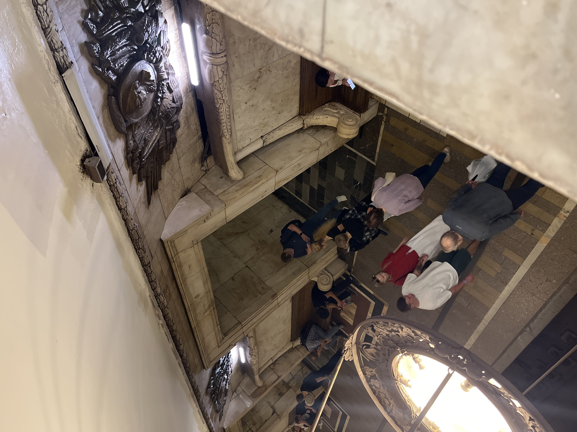 The image depicts an interior scene, likely a public space with ornate architectural details, possibly a metro station or historical building in Moscow, Russia. Several people are visible, some walking and others standing, many looking at their phones. The decor features marble cladding, intricate metalwork, and decorative plaster or stucco elements on the ceiling and walls. A large, decorative chandelier hangs in the foreground, illuminated. The overall lighting is somewhat dim, with artificial lights visible on the ceiling. The perspective is from above, looking down into the space.
