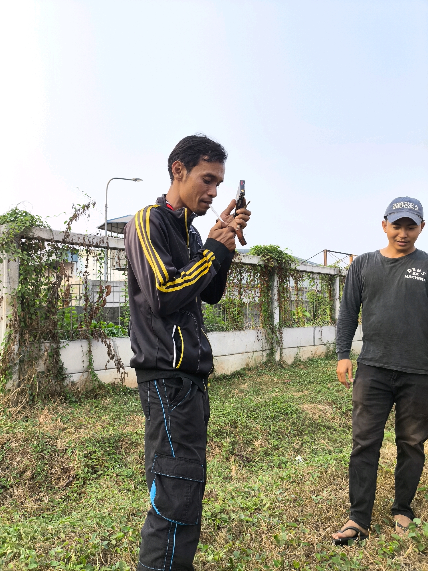 Two men are standing outdoors in a grassy area, likely in the city of Mekarwangi, Indonesia. The man on the left, wearing a black tracksuit with yellow stripes and black pants with blue trim, is holding a small object, possibly a lighter, to a cigarette and appears to be smoking. He is looking down at the cigarette. The man on the right, wearing a dark long-sleeved shirt with a graphic and black pants, stands to his right, facing forward with his hands by his sides. He is wearing a blue baseball cap with white lettering. Behind the men is a concrete fence overgrown with vines, a street lamp, and some industrial-looking structures in the distance under a pale sky. The ground is covered with dry grass and some green vegetation.