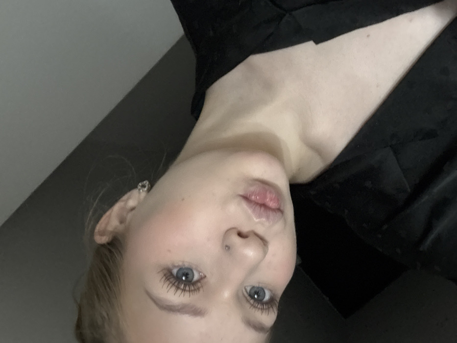 The image features a close-up, angled selfie of a young adult female. She has light-colored hair, blue eyes with enhanced eyelashes, rosy cheeks, and full lips puckered into a "kissing" expression. A small silver stud piercing is visible on her philtrum, and a decorative stud earring is present in her left earlobe. She is wearing a black garment, possibly a jacket or robe, with a subtle textured pattern. Her head is tilted to the left, and she is looking directly at the camera. The background is a uniformly dark gray, consistent with an indoor environment. The location is an indoor setting in the city of Yekaterinburg, Russia.