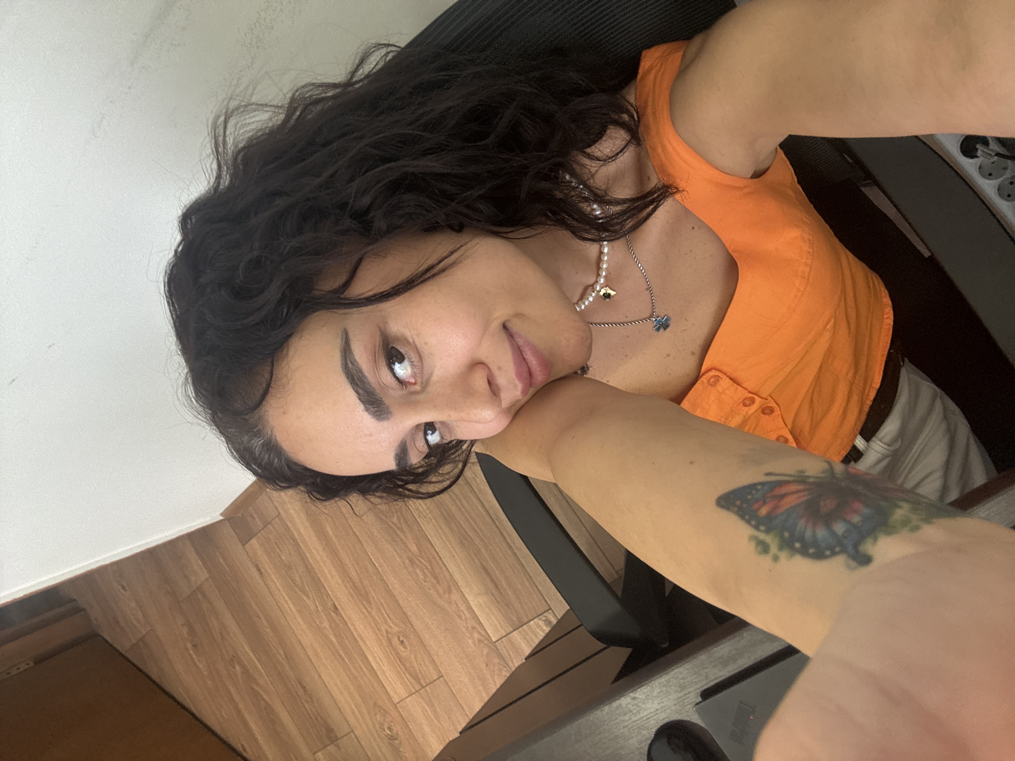 A woman with dark, curly hair and a tattoo of a butterfly on her left arm is sitting in a black office chair in front of a desk. She is wearing an orange top and a pearl necklace with a small blue pendant. The background shows a wooden floor and a white wall, suggesting an indoor setting. The image appears to have been taken in Tbilisi, Georgia.