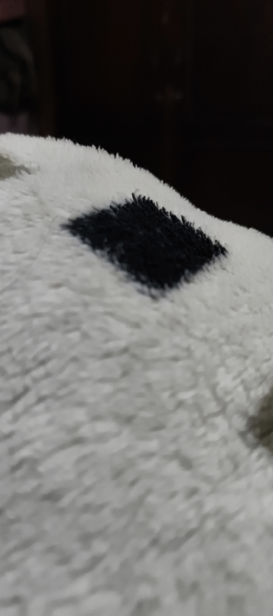 A close-up, low-angle shot shows a black diamond pattern on a fluffy white surface. The texture is soft and appears to be fabric, possibly a blanket or rug. The background is dark and out of focus, suggesting a dimly lit indoor environment. There is a single light-colored hair strand extending from the white fabric, to the left of the black diamond. The scene is abstract, with no identifiable objects or people beyond the patterned fabric itself. The image is likely from Muaro Jambi, Indonesia, though the visual elements do not provide specific clues to this location.