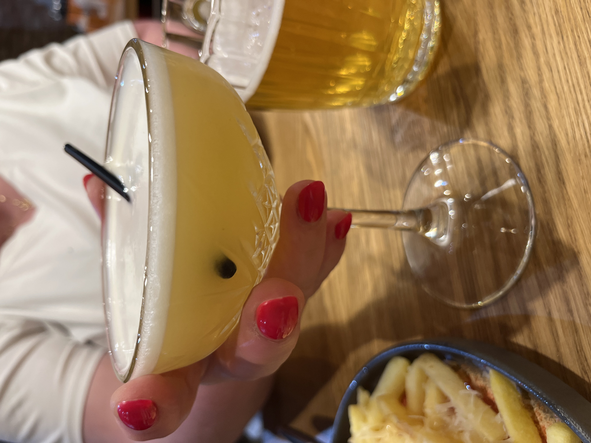 In Ново-Никольское, Russia, a close-up shot captures a hand with bright red nail polish holding a coupe glass filled with a frothy, yellow cocktail. A dark straw is submerged in the drink, and a single passion fruit seed is visible. To the right of the cocktail, a pint glass of golden beer is partially visible. In the lower left corner, a dark bowl contains what appears to be french fries topped with shredded cheese, some of which have fallen onto a red base. The scene is set on a wooden table, illuminated by soft lighting that creates subtle reflections on the glassware and table surface. The background shows a person wearing a white top.
