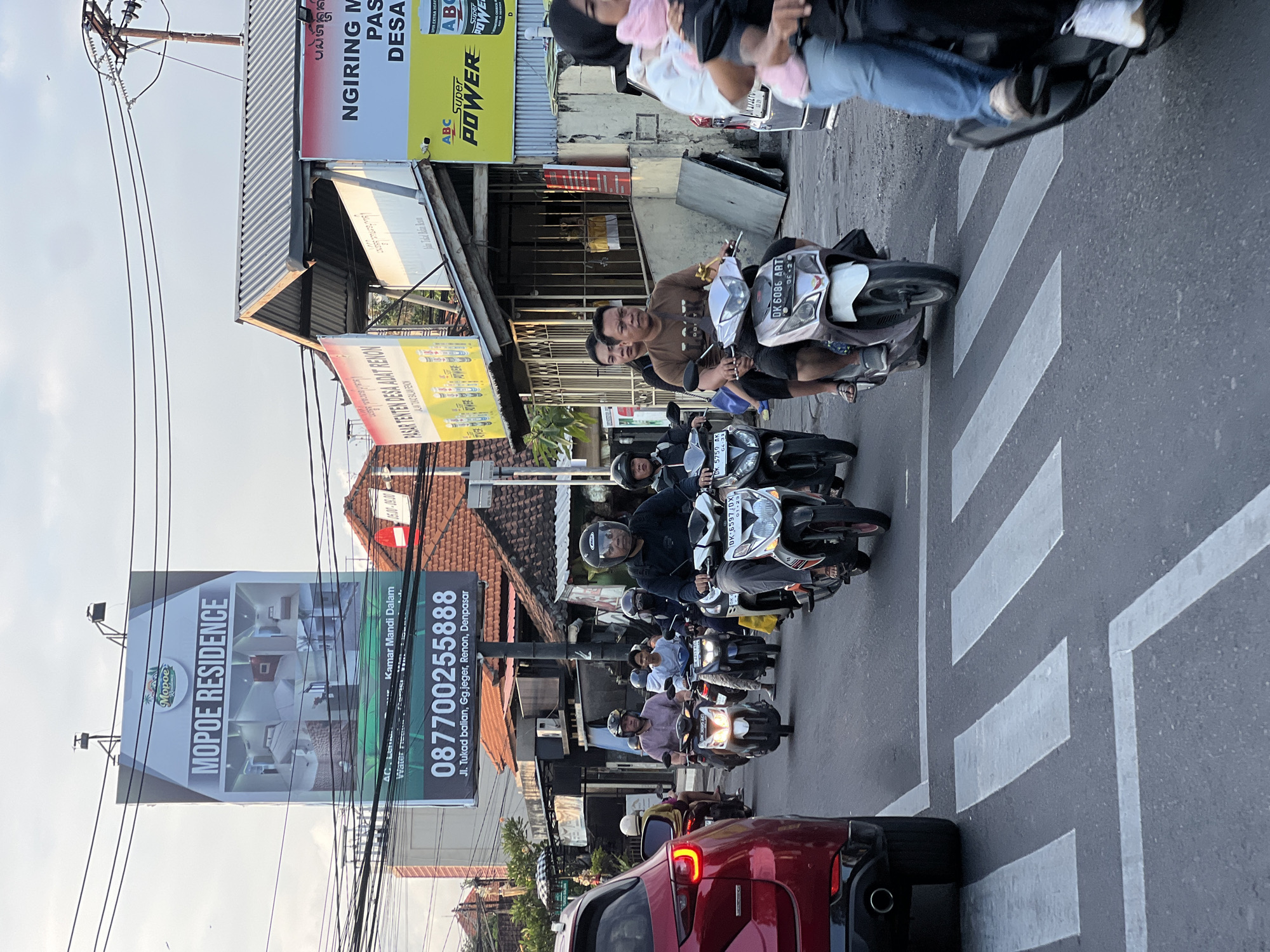 The image captures a street scene in Denpasar, Indonesia, likely during daytime, characterized by a moderate level of traffic, primarily consisting of motorcycles. In the foreground, a red SUV is positioned, partially obscuring a crosswalk. Several individuals are riding motorcycles, navigating the road. Prominently displayed on a billboard in the background is an advertisement for "MOPOE RESIDENCE" with contact information and an address on "Jl. Tukad balian, Gg.jeger, Renon, Denpasar". Other commercial advertisements are visible on buildings, including one for "ABC Super POWER". The environment appears to be an urban setting with buildings, utility poles, and power lines lining the street. The sky suggests it is late afternoon or early evening with a light haze.