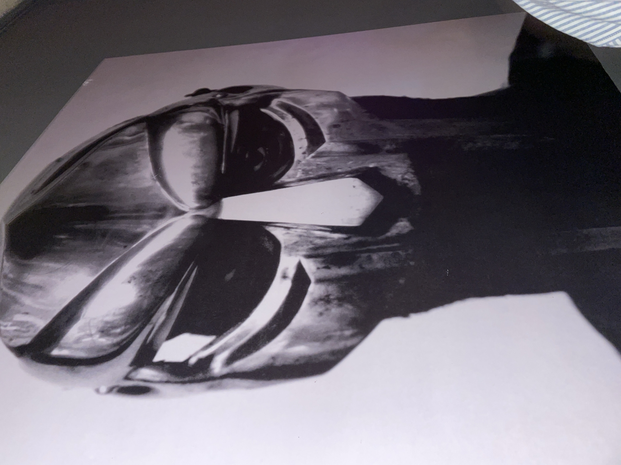 The scene captures a large black and white print of a stylized metallic mask, characteristic of the rapper MF DOOM, displayed prominently on a wall in Barcelona, Spain. The mask, which dominates the frame, is rendered with strong contrasts, highlighting its angular features, including a heavily defined brow, narrow eye slits, and a distinctive pointed nose guard. Its surface appears textured and metallic, with light catching various facets and deep shadows forming beneath the chin and around the neck area. The print's background is a bright, uniform white or light grey, providing a stark contrast to the dark tones of the mask. The print itself is affixed to a dark grey wall, visible along the top right and right edges of the frame. In the lower right corner, a portion of a person's arm and shoulder, wearing a blue and white striped shirt, is visible, indicating the presence of an observer taking the photograph.