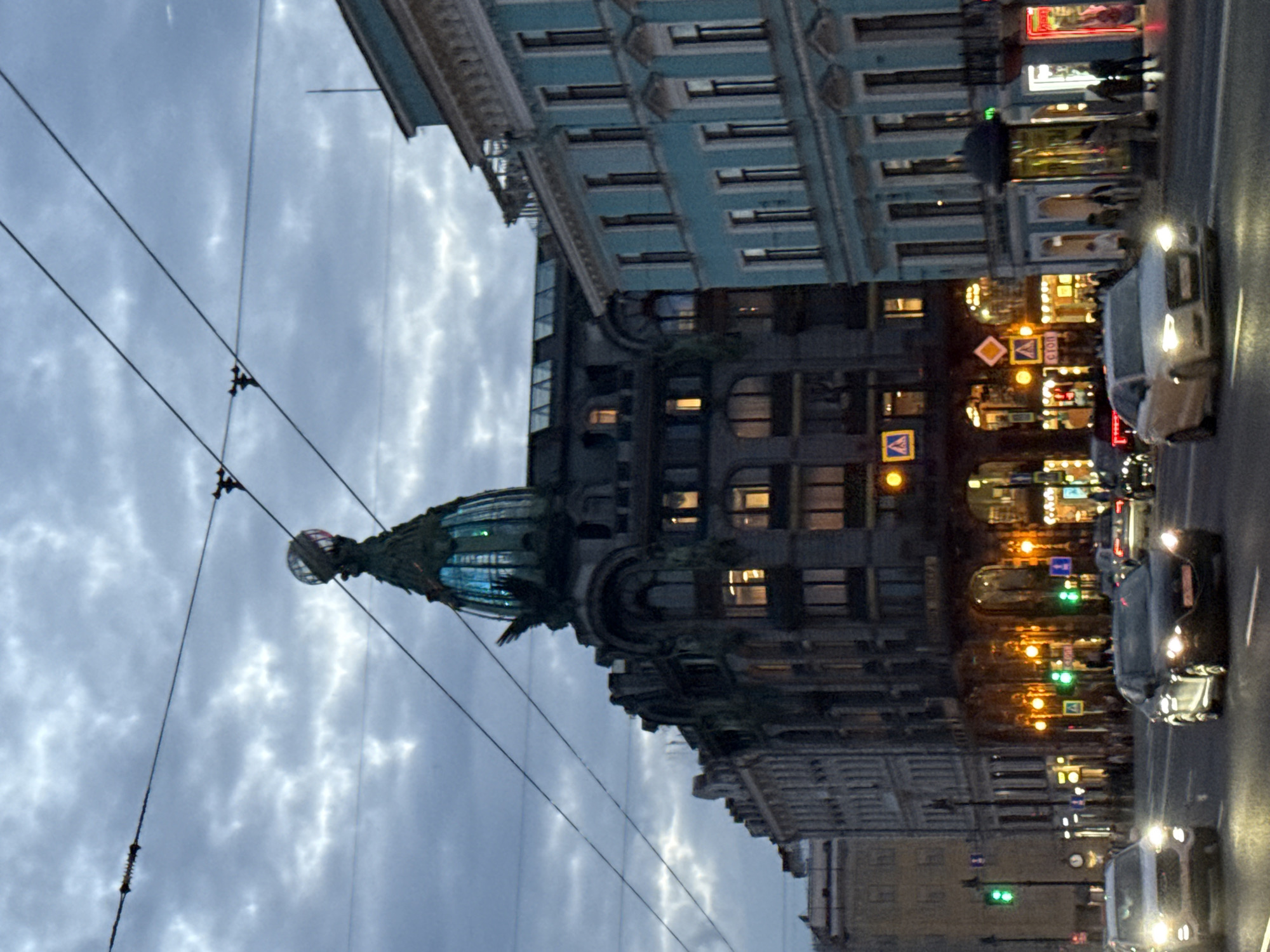 The image depicts Nevsky Prospekt in Saint Petersburg, Russia, at dusk. The iconic Singer House building, with its distinctive glass dome topped by a globe and eagle sculpture, dominates the center of the frame. The sky is overcast with dramatic clouds.
Traffic is visible on the multi-lane street, with cars driving in both directions. Headlights illuminate the road and the surrounding buildings. Pedestrians can be seen on the sidewalks, and shop windows glow with interior lighting. The architecture of the surrounding buildings is typical of Saint Petersburg, with ornate facades and multi-story structures. Overhead wires, likely for trams or trolleybuses, crisscross the sky. The overall atmosphere is urban and slightly moody, characteristic of a city at twilight with active street life.