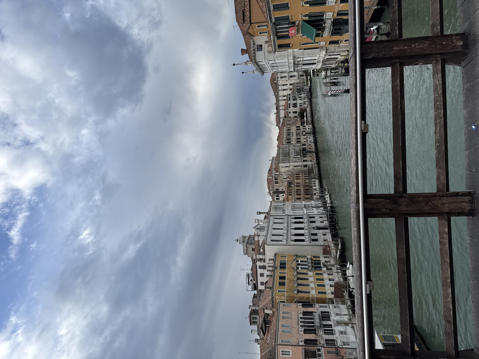 The image depicts a panoramic view of the Grand Canal in Venice, Italy, observed from a wooden bridge railing positioned in the foreground.
The sky occupies the upper two-thirds of the frame, characterized by dense, dark grey, overcast clouds, with some lighter patches revealing traces of blue sky towards the top and right. The light suggests daytime conditions.
The Grand Canal, a significant waterway, dominates the midground. Its water appears a murky greenish-blue, showing minimal surface disturbance. Numerous dark wooden mooring poles (bricole) are sparsely distributed within the canal and along its banks. Several small, white-hulled boats are moored to these poles or directly against the buildings along the left bank.
Both banks of the canal are lined with a continuous succession of multi-story historic buildings exhibiting Venetian architecture. On the left bank, the buildings present a variety of colors, including pale yellow, off-white, light pinkish-brown, and darker terracotta. Notable features include numerous rectangular and arched windows, balconies with black iron railings, and terracotta tiled roofs. A large, prominent white-faced building with multiple arched windows stands out on the left. Further down the canal on the left, a rounded dome or cupola is visible above the rooftops.
On the right bank, the buildings also display varying hues of orange, white, and reddish-brown. One building in the mid-right features a red banner or sign and a green awning over a lower-level balcony. Another building further down on the right bank exhibits distinctive twin spires on its roofline. The architecture generally comprises stone or stucco facades with intricate details.
In the immediate foreground, a dark brown wooden bridge railing spans the width of the image. It consists of two vertical posts supporting three horizontal planks, with an additional silver-colored metal bar attached to the uppermost horizontal plank. A small, indistinct blue object rests on the wooden bridge deck beneath the railing, slightly to the left of the central vertical post. No individuals or discernible actions are present within the frame.