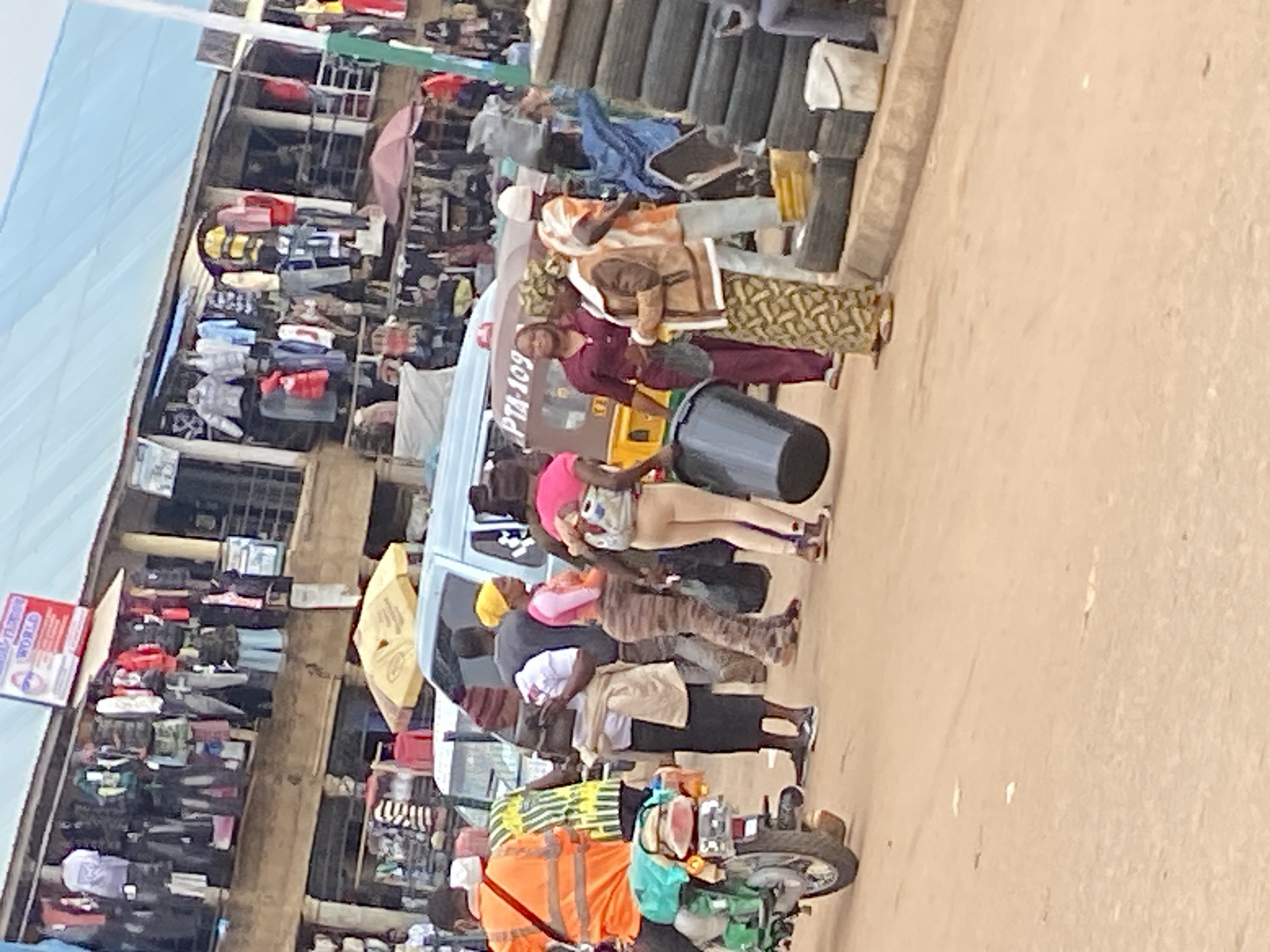 In Bukuru, Nigeria, a bustling marketplace scene unfolds. A man in an orange reflective vest rides a green motorcycle, its rear tire visible in the foreground. He is accompanied by a person in a yellow and green striped shirt. Behind them, a group of individuals navigate the dusty street. One woman, wearing a pink top and patterned leggings, carries a child on her back, while another woman in a purple shirt holds a black bucket. A woman in a brown jacket with a patterned skirt also carries a black bucket.
In the background, a light blue minivan with "PTA-109" visible on its side is parked, suggesting transportation services. Shops line the street, displaying a variety of clothing items hung from poles and shelves, including shirts, jackets, and pants. Several umbrellas, one yellow and one red, offer shade in the market. A stack of used tires is visible on the right side of the image, next to a white bucket. The ground is a dry, reddish-brown dirt. The overall atmosphere is one of vibrant commercial activity and daily life in a Nigerian town.