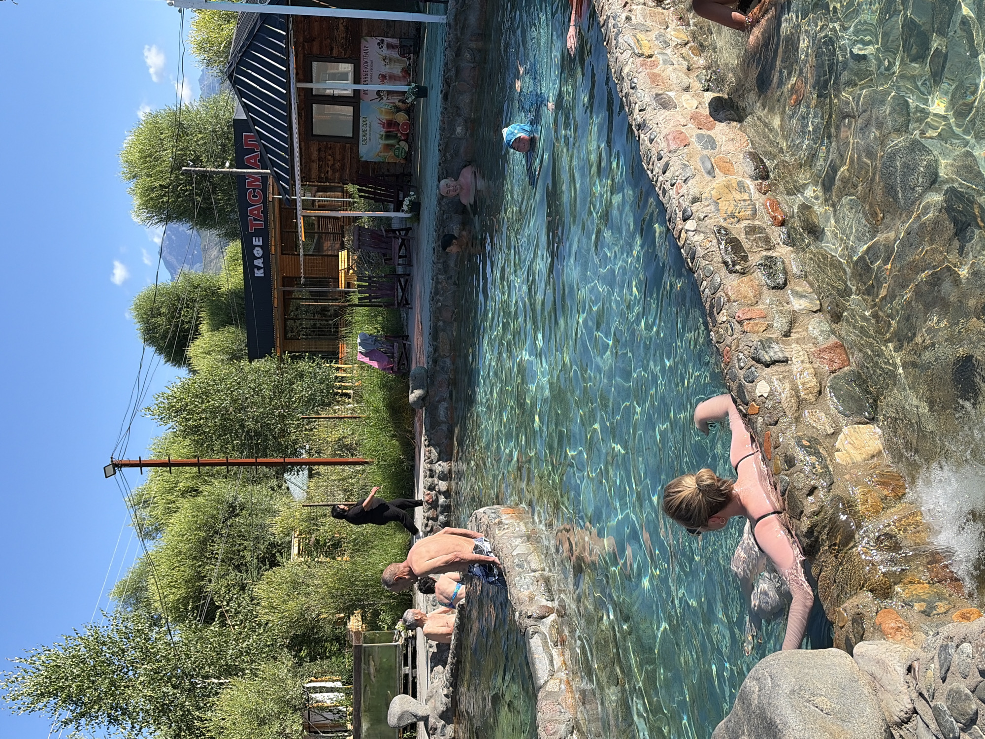 An outdoor hot spring resort is depicted on a clear, sunny day, with a bright blue sky and fluffy white clouds. Several people are visible enjoying the mineral-rich, turquoise water. In the foreground, a woman with blonde hair tied up in a bun is relaxing in the water, leaning back against the stone edge of the pool. The pool itself is partially bordered by a rustic stone wall. Further into the pool, other individuals are swimming and wading. In the background, there is a wooden building with a sign that reads "КАФЕ ТАСМАЛ" (Cafe Tasmaal), suggesting a cafe or restaurant facility. Trees and lush green foliage surround the area, and in the distance, mountains are visible under the bright sky. The overall scene conveys a sense of leisure and relaxation in a natural, resort-like setting.