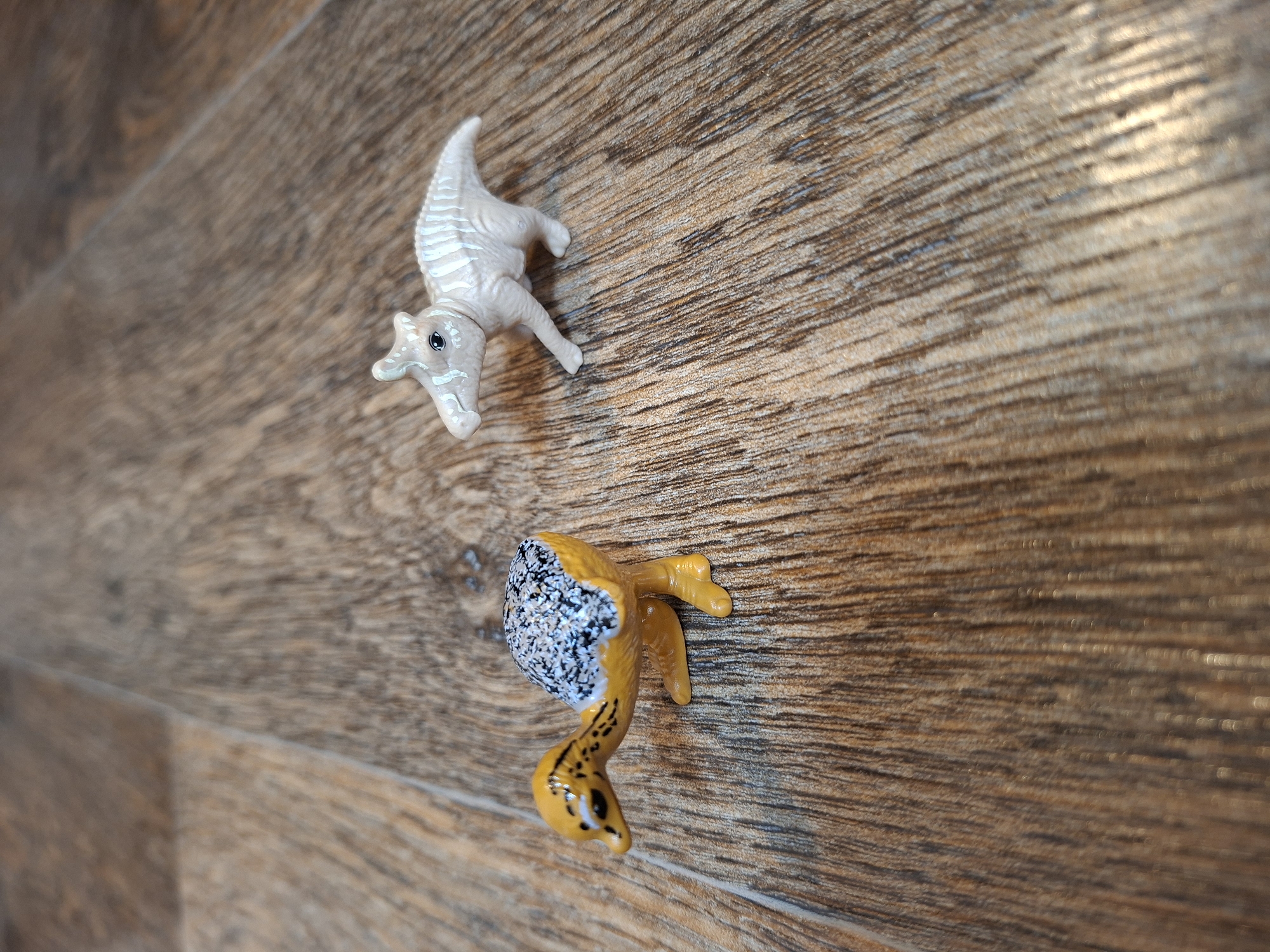 Two small, plastic dinosaur figurines are placed on a wooden surface with a wood grain pattern. The figurine on the left is yellow and orange with a speckled black and white back. It is depicted in a crouching position. The figurine on the right is a pale, off-white color with light blue stripes. It is standing on all fours and appears to be moving. The lighting in the image is soft, creating some shadow and highlights on the wooden surface. There are no people or other discernible objects in the scene. The setting does not provide any specific location information for Gorkovskiy, Russia.