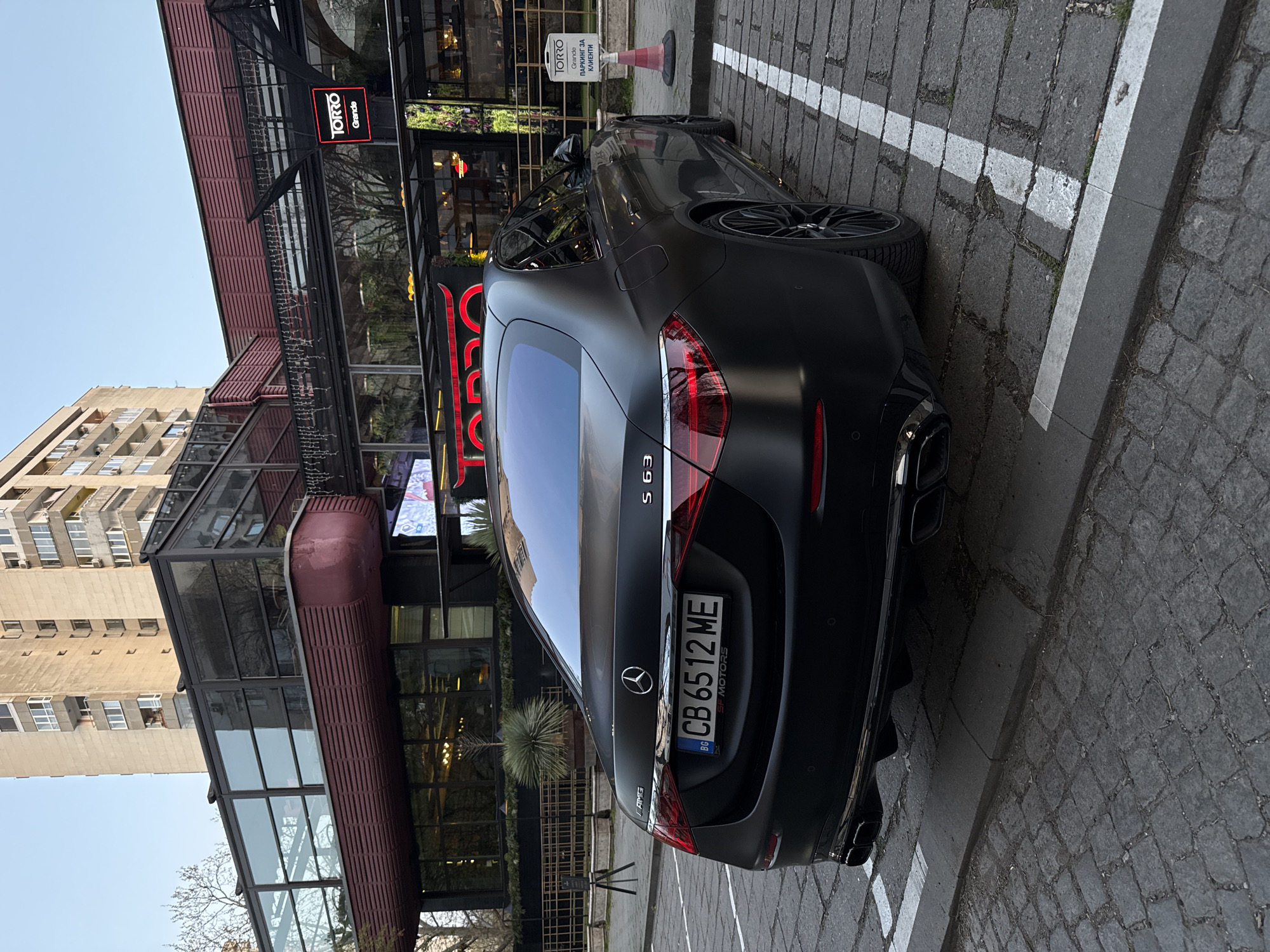 The image shows a matte black Mercedes-AMG S 63 parked on a brick-paved street next to the TORRO Grande restaurant in Plovdiv, Bulgaria. The car is parked in a designated spot marked by a white line, with its rear facing the camera. The license plate number is CB 6512 ME. A sign indicating "TORRO Grande Паркинг за клиенти" (Parking for clients) is visible next to a small orange traffic cone. The restaurant's facade is modern with glass and steel construction, and a multi-story residential building is also visible.