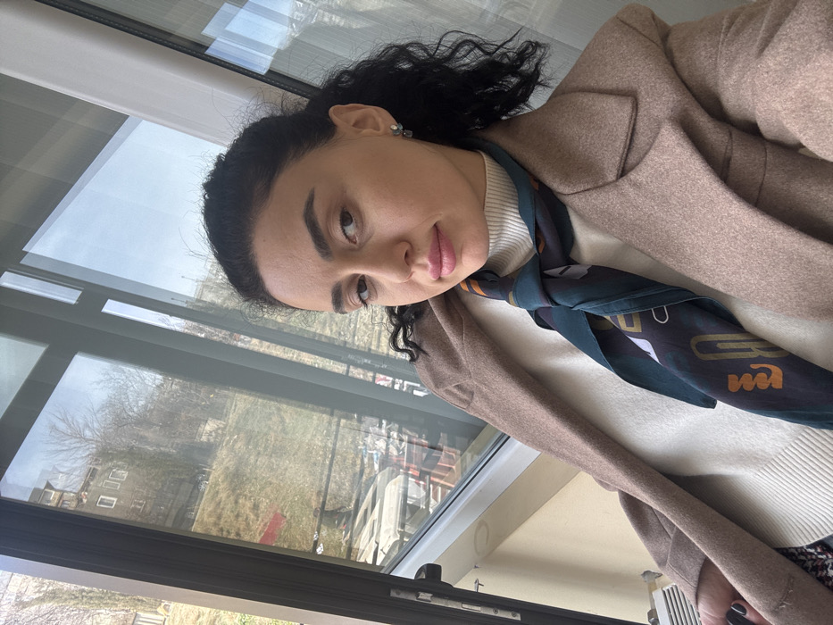 A female subject, estimated to be between 25 and 40 years old, is depicted in a medium shot, looking directly at the camera. She has dark, curly hair, dark eyebrows, light brown eyes, and is wearing light makeup with pink lipstick. Her attire includes a light brown, possibly wool, overcoat worn over a white ribbed turtleneck sweater. A dark teal silk scarf with abstract geometric patterns in orange, white, and black is tied around her neck. She wears small, multi-stone stud earrings. The subject's left hand, with dark nail polish, is partially visible on the lower left.
The subject is positioned indoors, in front of a large, dark-framed window or glass door. Through the window, an outdoor scene is visible under a bright sky. The background shows a sloped, grassy hillside with bare deciduous trees, indicative of late autumn or winter. In the middle distance, a light-colored multi-story building with several windows is visible on the hillside. Below the window on the left, a white radiator or heating unit is present. The general environment suggests a residential setting overlooking a hilly urban landscape. Based on the hilly terrain and architectural cues, the location is consistent with a residential area in Tbilisi, Georgia.