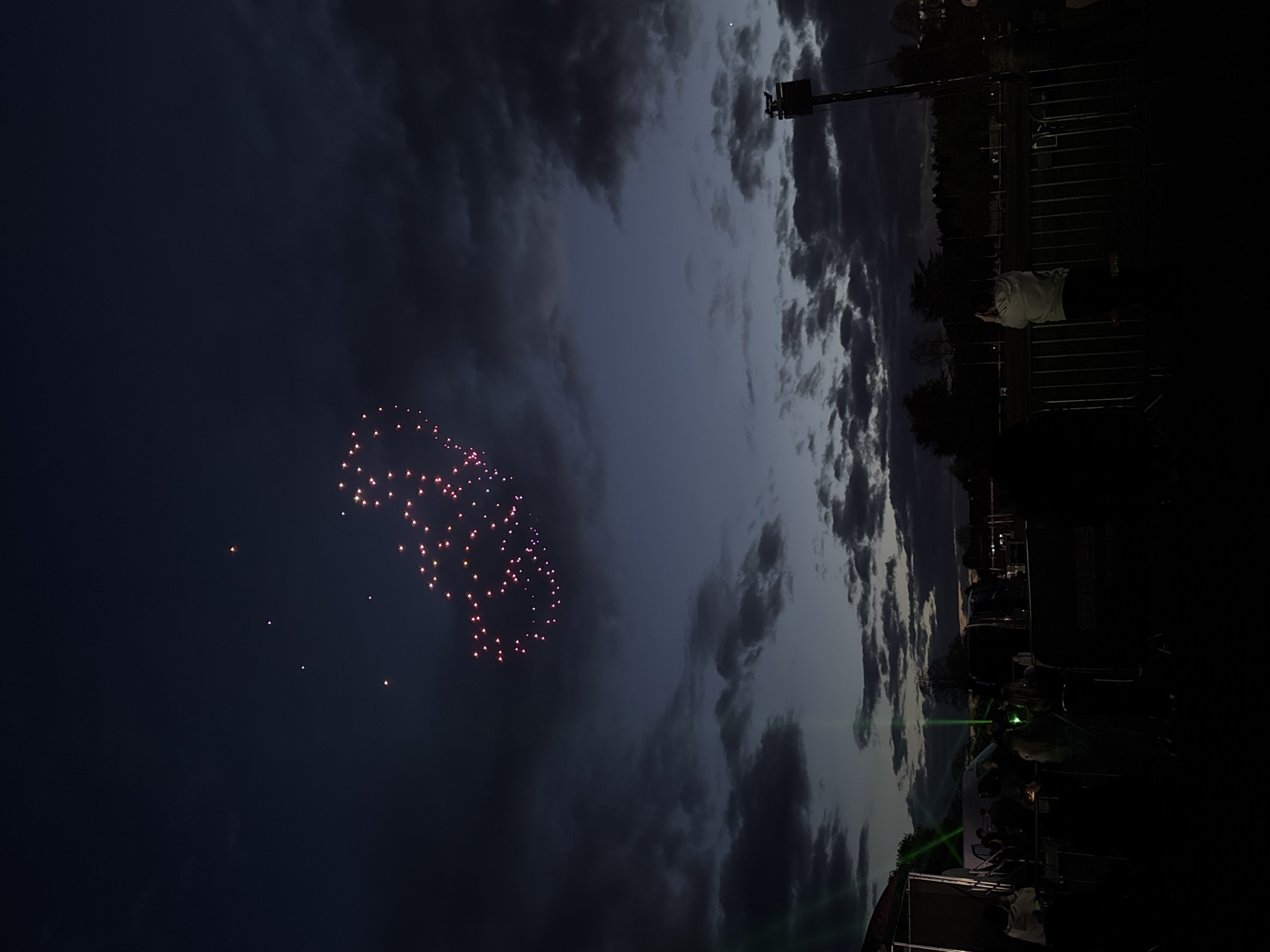 The image depicts a night scene in South Norfolk, United Kingdom. A firework display is visible in the dark sky, forming a pattern with pinkish-red bursts. The sky is also partially obscured by clouds. A tall structure, possibly a light tower, stands near the horizon. In the foreground, a crowd of people is present, with some individuals near a barrier or fence. Green lights illuminate part of the scene.