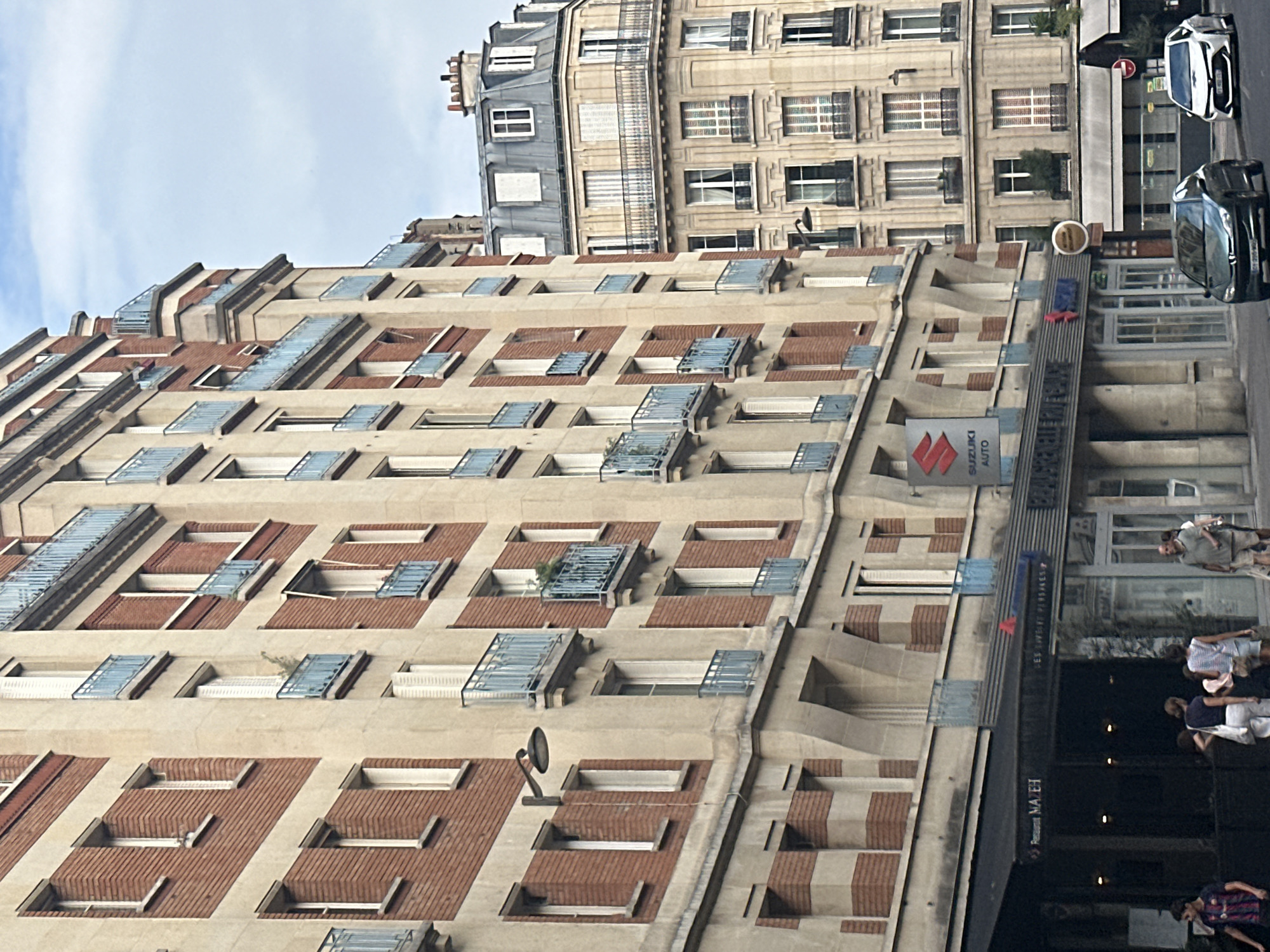 The image shows a street scene in Paris, France, with buildings lining a road. The buildings are made of brick and stone, with multiple stories and balconies. One building on the left has a red brick facade with cream-colored horizontal bands. Above a storefront, a sign for "SUZUKI AUTO" is visible. The street level has storefronts, including one with a black awning. Several people are walking on the sidewalk, and two cars are parked on the street. The architecture suggests an urban environment in Paris.