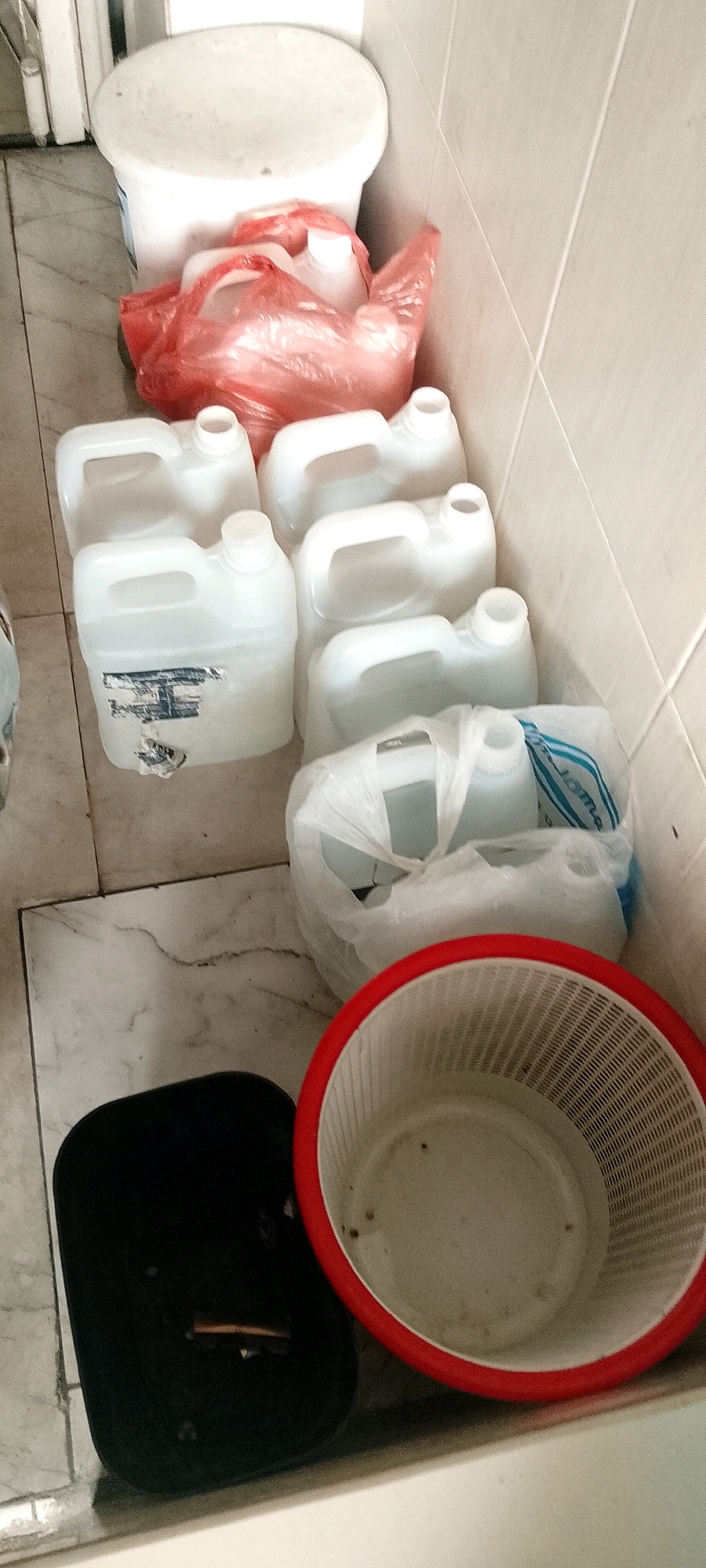 The image shows a corner of a room, likely a bathroom or utility area, with tiled walls and floor. In the foreground, a black rectangular object rests on the floor, with a small brown object on top. To its right is a white, mesh-like basket with a red rim, containing some dirt or debris. Behind these items, a line of white plastic jugs with handles and white caps is arranged. Some of these jugs are partially obscured by a red plastic bag filled with more white plastic containers. The jugs appear to be empty or partially filled with a clear liquid. In the background, a large white cylindrical container stands against the wall. The lighting is artificial and appears to be coming from above. The overall impression is one of storage or preparation for some task.