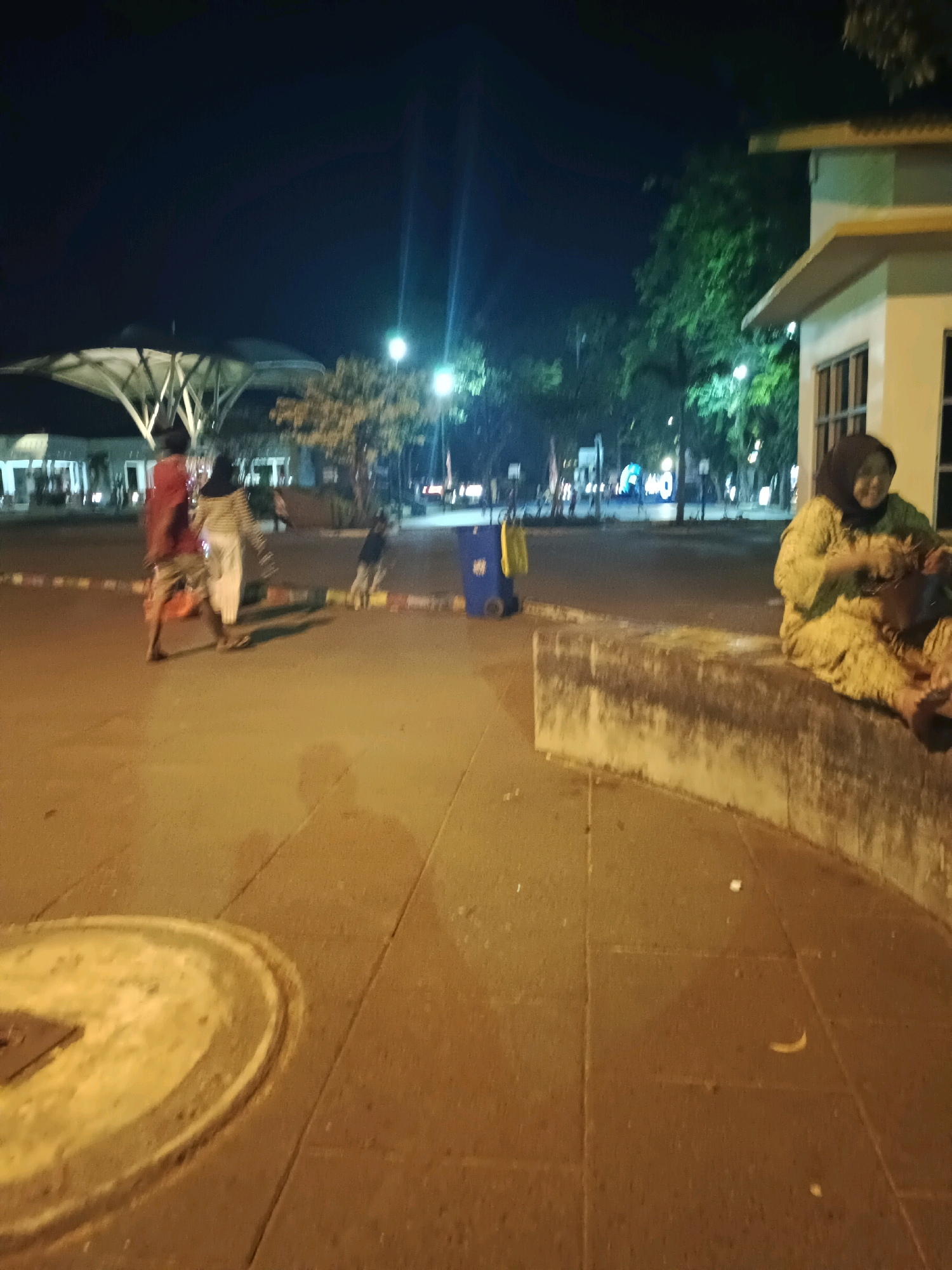 The image depicts an outdoor scene at night, likely in a public area in Lamongan, Indonesia. In the foreground, a concrete sidewalk and curb are visible, with a circular metal grate embedded in the pavement. To the right, a woman wearing a yellow patterned outfit and a hijab is seated on the concrete curb, holding a dark bag. She appears to be looking down at the bag.
In the middle ground, two individuals are walking across the pavement, one in a red top and shorts, and the other in a white and striped outfit with a head covering. Behind them, a blue wheeled bin is visible. The background shows a street with trees, lighting, and the facade of a building on the right. A covered structure with a white canopy supported by visible beams is present on the left. The overall lighting suggests artificial illumination from streetlights and building lights.