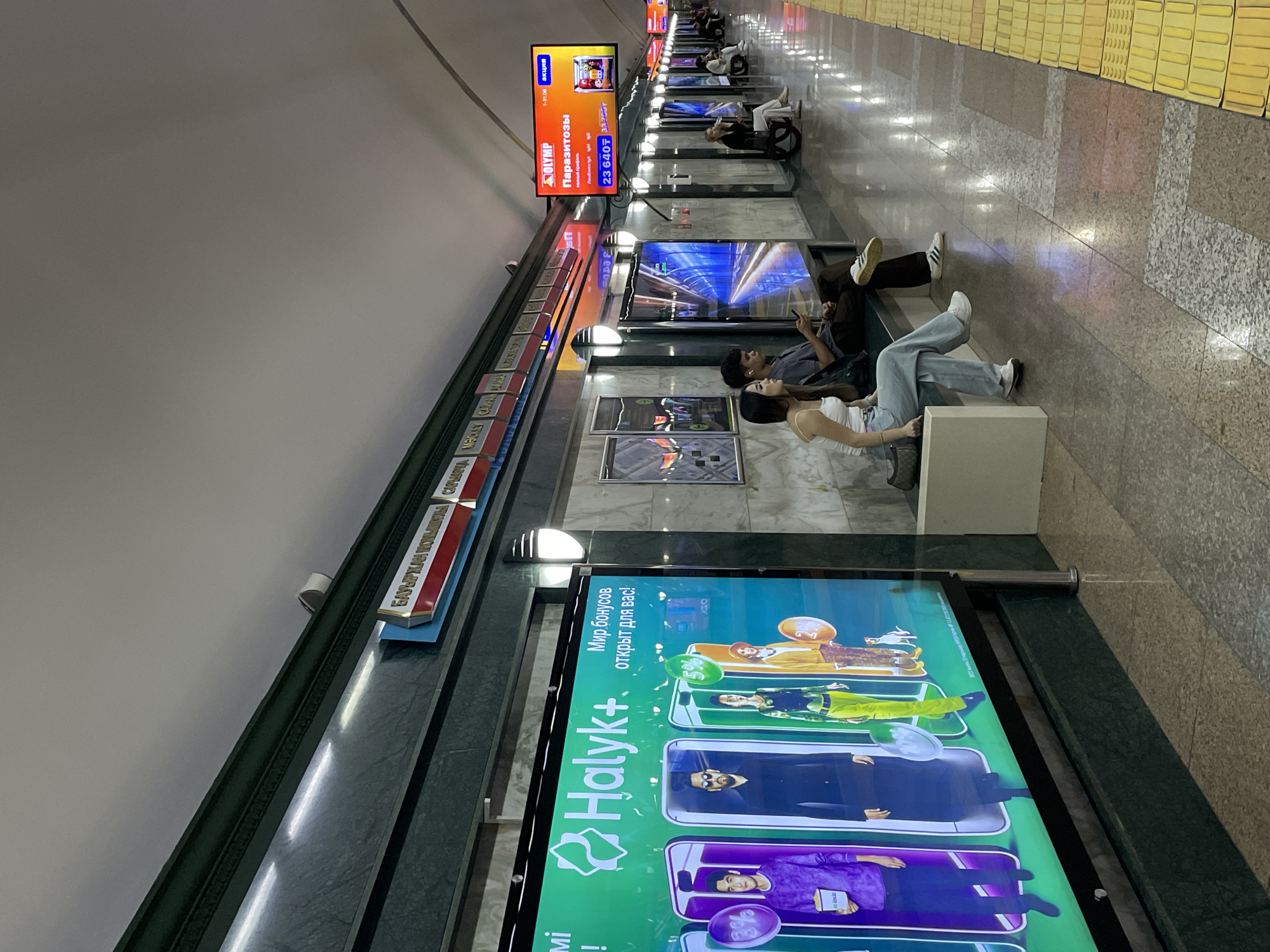 The image depicts a metro station platform in Almaty, Kazakhstan. A large digital advertisement for "Halyk+" is prominently displayed on the left, featuring people in stylized clothing and mobile phone graphics. To the right, a smaller digital advertisement for "OLYMP" advertises "Паразитозы" (Parasitoses) with a price.
Several individuals are visible on the platform. In the foreground, a young couple sits on a bench, with the woman looking to her left and the man looking down at his phone. Further down the platform, other passengers are seated or walking. The platform itself is constructed with polished stone flooring, featuring a tactile warning strip along the edge. Overhead, a green decorative border runs along the wall, punctuated by lighting fixtures. The station's signage is visible at the top, with text that appears to be in Cyrillic script. The overall environment is clean and modern, typical of an urban metro system.