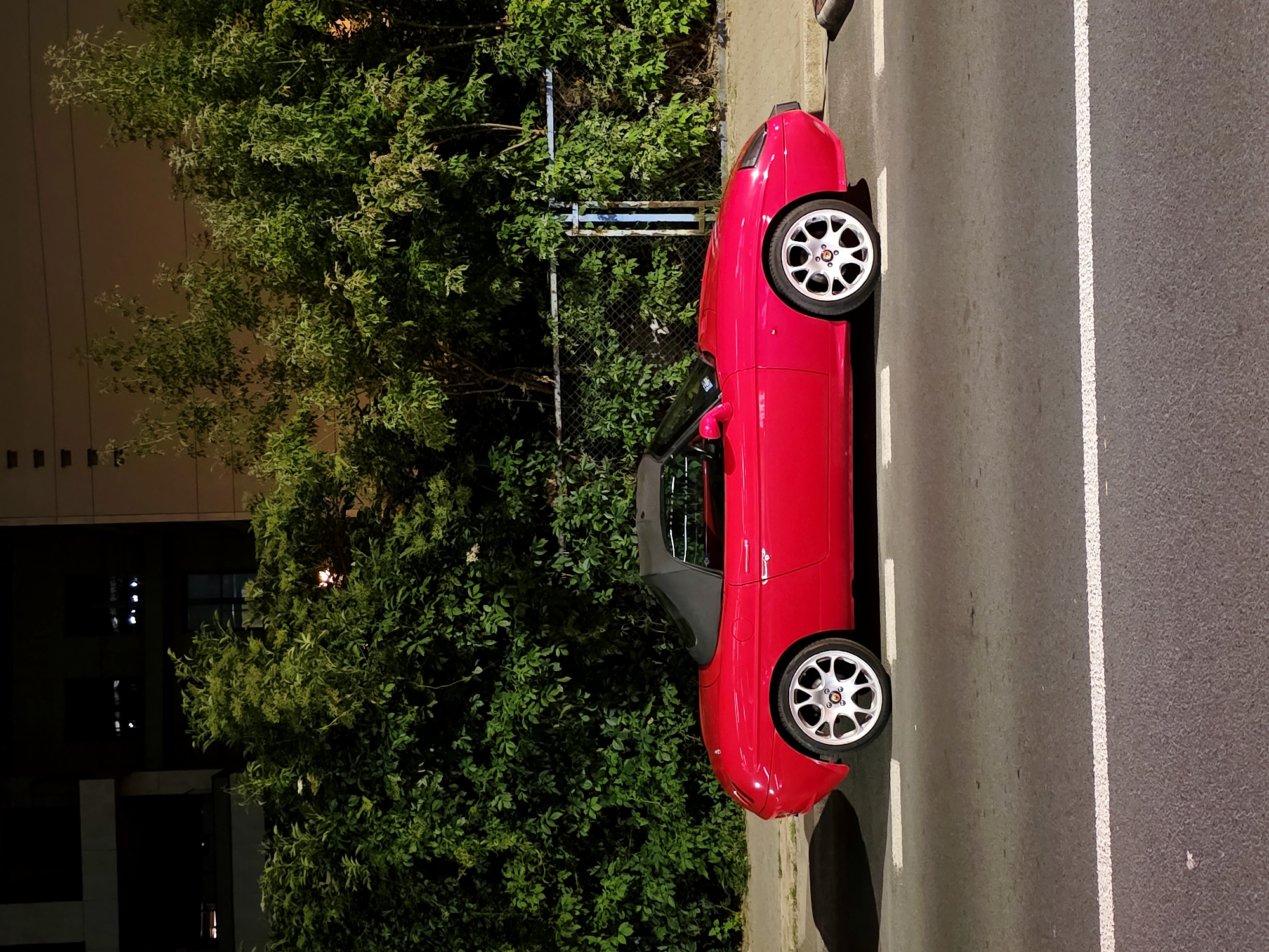 A red convertible sports car, possibly a Fiat Barchetta, is parked on the side of a street at night. The car is facing left in the frame. The street has white lane markings. Behind the car is a dense green hedge, and beyond that, a chain-link fence with blue posts. In the background, there are buildings and trees. The lighting suggests an urban environment at night. There are no people visible in the image. The scene is located in Warsaw, Poland.