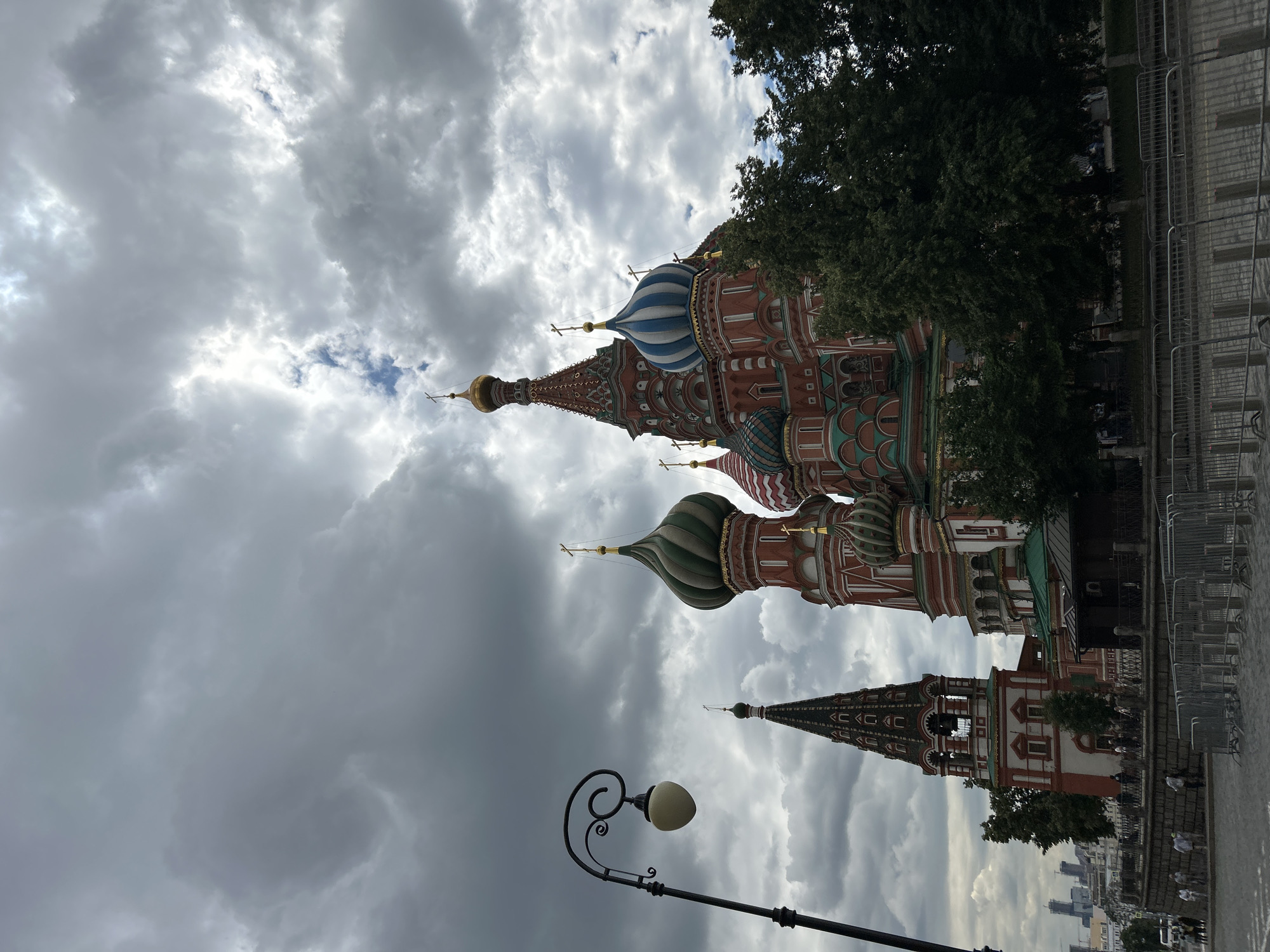 The image depicts Saint Basil's Cathedral in Moscow, Russia, under a cloudy sky. The cathedral's iconic colorful onion domes are visible, along with a tall, slender bell tower. In the foreground, a black lamppost with a decorative scroll arm and an ovular lamp is positioned to the left. A stone wall and metal barriers separate the paved ground from the area in front of the cathedral. Several trees with dense green foliage are situated between the viewer and the cathedral. In the background, a cityscape with tall buildings can be seen. A few people are present on the paved area, walking and standing. The sky is filled with dark, dramatic clouds, suggesting overcast weather.