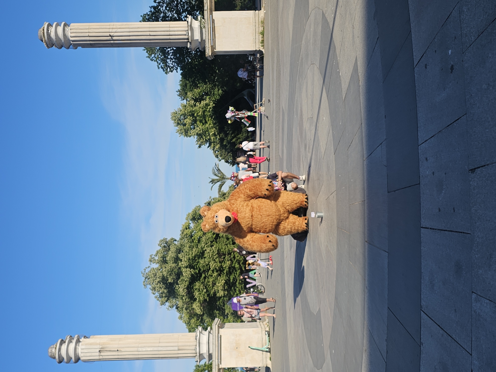 A person in a furry brown bear costume with a red bow around its neck is standing in an outdoor plaza. The bear costume is large and appears to be made of plush material. The person inside the costume is wearing dark shoes. In front of the bear, on the ground, is a white disposable cup. The plaza is paved with large, gray square tiles. There is a large circular pattern visible in the paving stones behind the bear.
In the background, several other people are visible, some walking and some standing. Among them, a person is selling colorful balloons and other small toys from a cart. Trees with lush green foliage are present in the mid-ground and background, suggesting it is a warm season.
Two large, fluted white columns with Corinthian capitals are visible in the foreground and mid-ground on either side of the frame, indicating a monumental or classical architectural setting. The sky above is a clear, bright blue, with a few wispy clouds. The sunlight suggests it is daytime, likely midday or afternoon, given the shadows. The overall mood seems light and perhaps festive, with the costumed character potentially entertaining passersby. There is no visible text in the image.