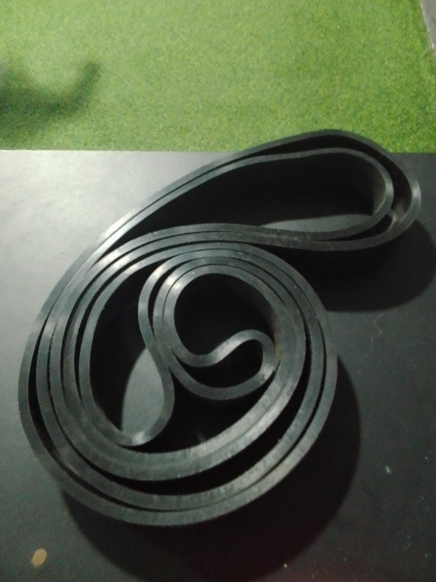 A black, rubber resistance band is coiled on a dark, flat surface. The background behind the surface is a blurred green texture, possibly artificial turf. The image is taken from a slightly elevated perspective, with the band occupying the majority of the frame. The lighting casts a shadow of the band onto the surface. The location in Juan Griego, Venezuela, is not discernible from the visual information in the image.