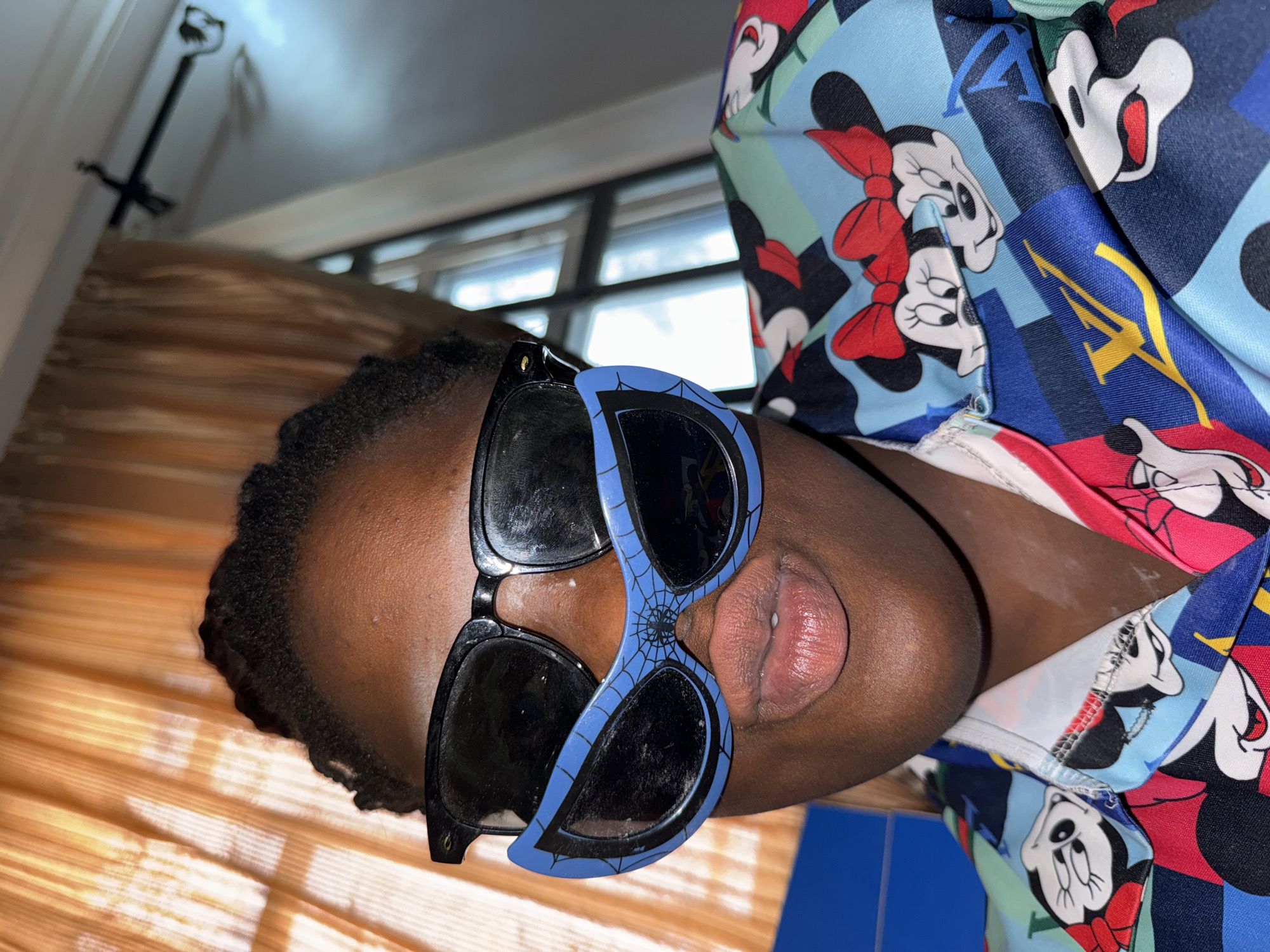The image shows a person wearing two pairs of sunglasses, one black and one blue with a spider web design. The person is wearing a shirt with a Mickey Mouse pattern. The background shows a window with blinds. It is difficult to confirm the location in Sabon Tasha, Nigeria, based on the image alone.