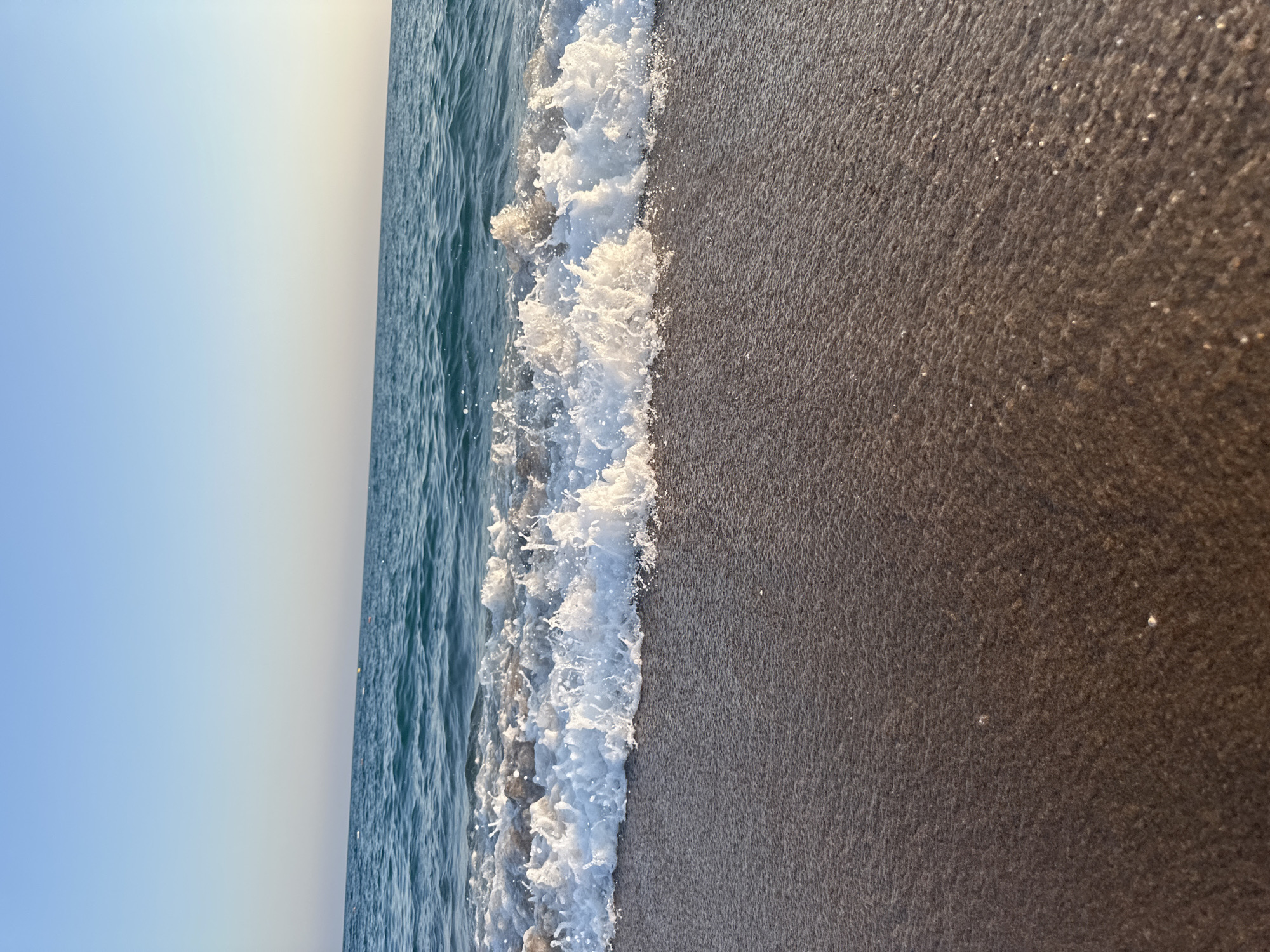 The image depicts a tranquil scene at the beach in Manavgat, Turkey. The foreground is dominated by wet, dark sand, reflecting the soft light of what appears to be either sunrise or sunset. A gentle wave is breaking, creating a foamy, white crest that washes over the sand. The ocean water is a deep blue-green, calm and rippling. The horizon is a clear demarcation between the sea and a pale blue sky, with a subtle gradient towards a lighter hue near the horizon, suggesting the time of day. No people or man-made objects are visible in this particular frame, emphasizing the natural beauty of the coastal environment.