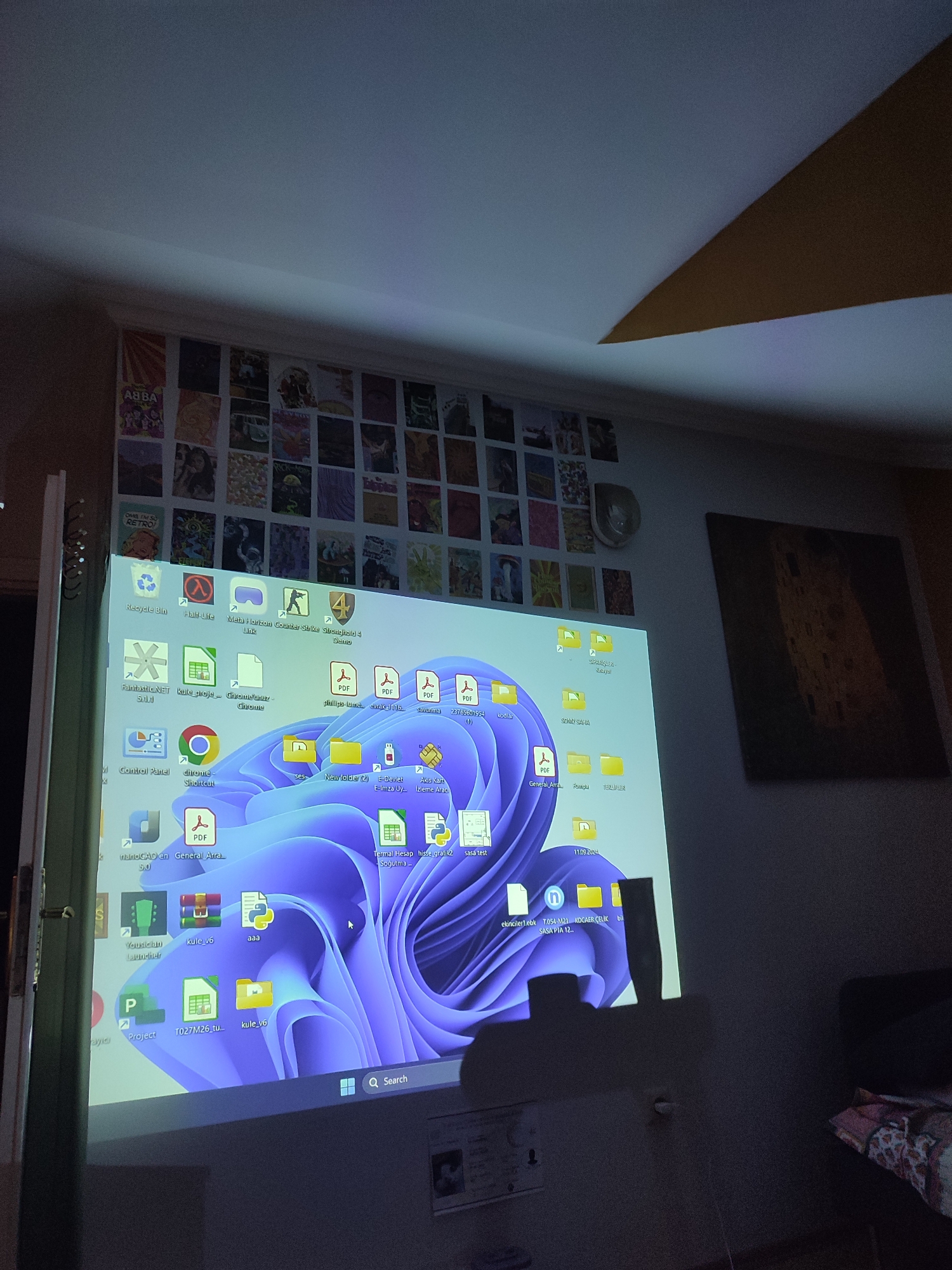A projector screen displays a Windows 11 desktop with numerous icons and folders. On the wall behind the projector screen, there is a collage of small posters or images. To the right of the projector screen, a large framed artwork hangs on the wall. The room is dimly lit, with a projector visible in the foreground casting the desktop image onto the wall. The image does not contain any elements that indicate it is located in Üsküdar, Turkey.