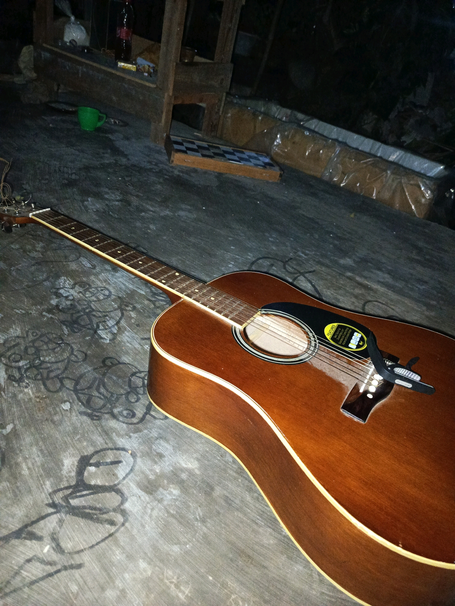 An acoustic guitar with a brown wood finish lies on a concrete surface. The guitar has a black pickguard and a black pickup attached to its soundhole. In the background, there is a wooden structure, a green cup, and some plastic-wrapped items. The scene appears to be outdoors, possibly in a yard or an alleyway, with some graffiti or chalk drawings on the concrete surface. The lighting suggests it is nighttime or a dimly lit area.