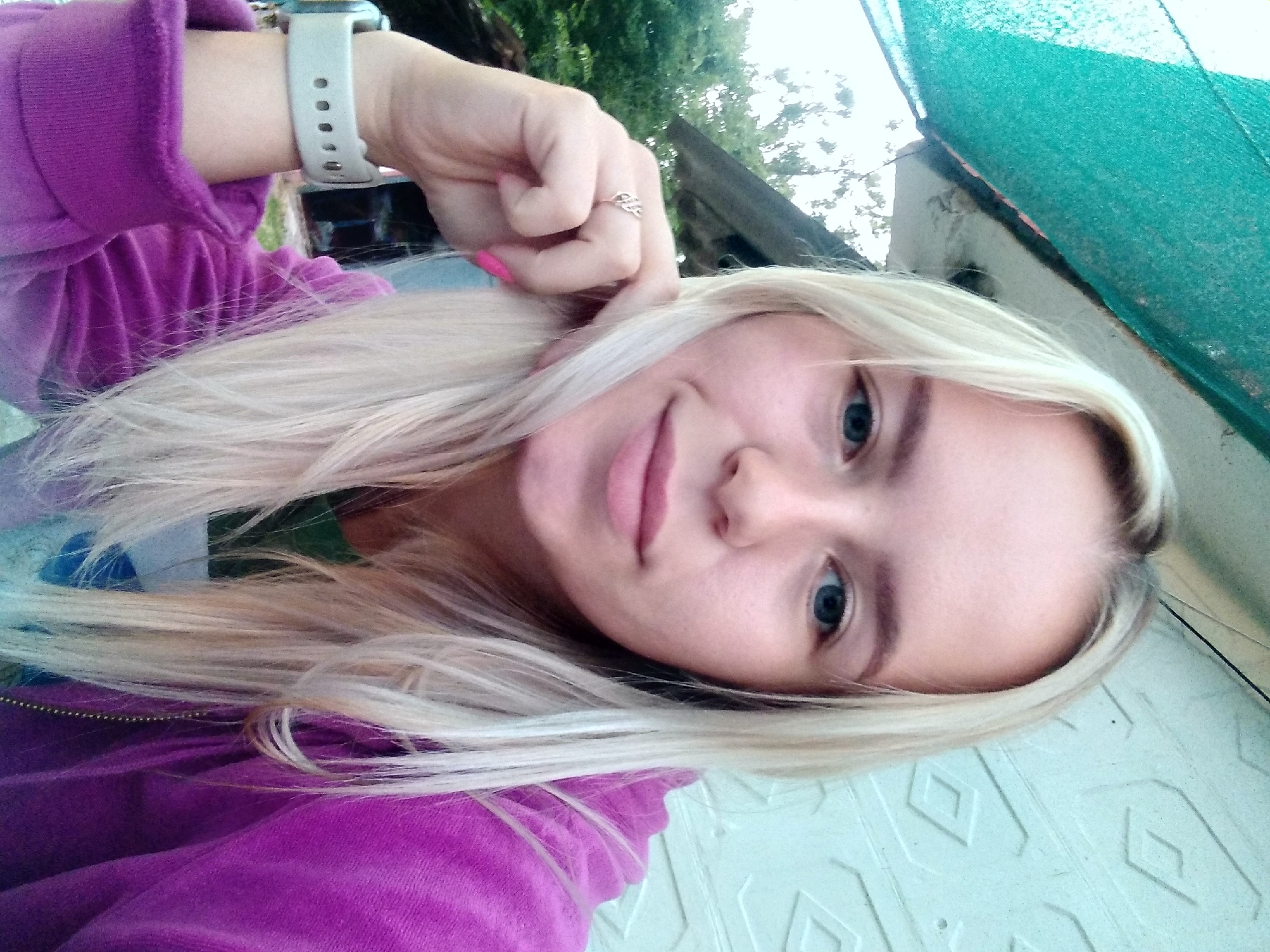 A young woman with blonde hair and blue eyes is pictured in an outdoor setting. She is wearing a purple hooded sweatshirt and a white watch on her left wrist. Her right hand is near her face, and she is wearing a ring on her right ring finger. Behind her, a textured wall and some greenery are visible. The scene appears to be in the city of Radisne, Ukraine.