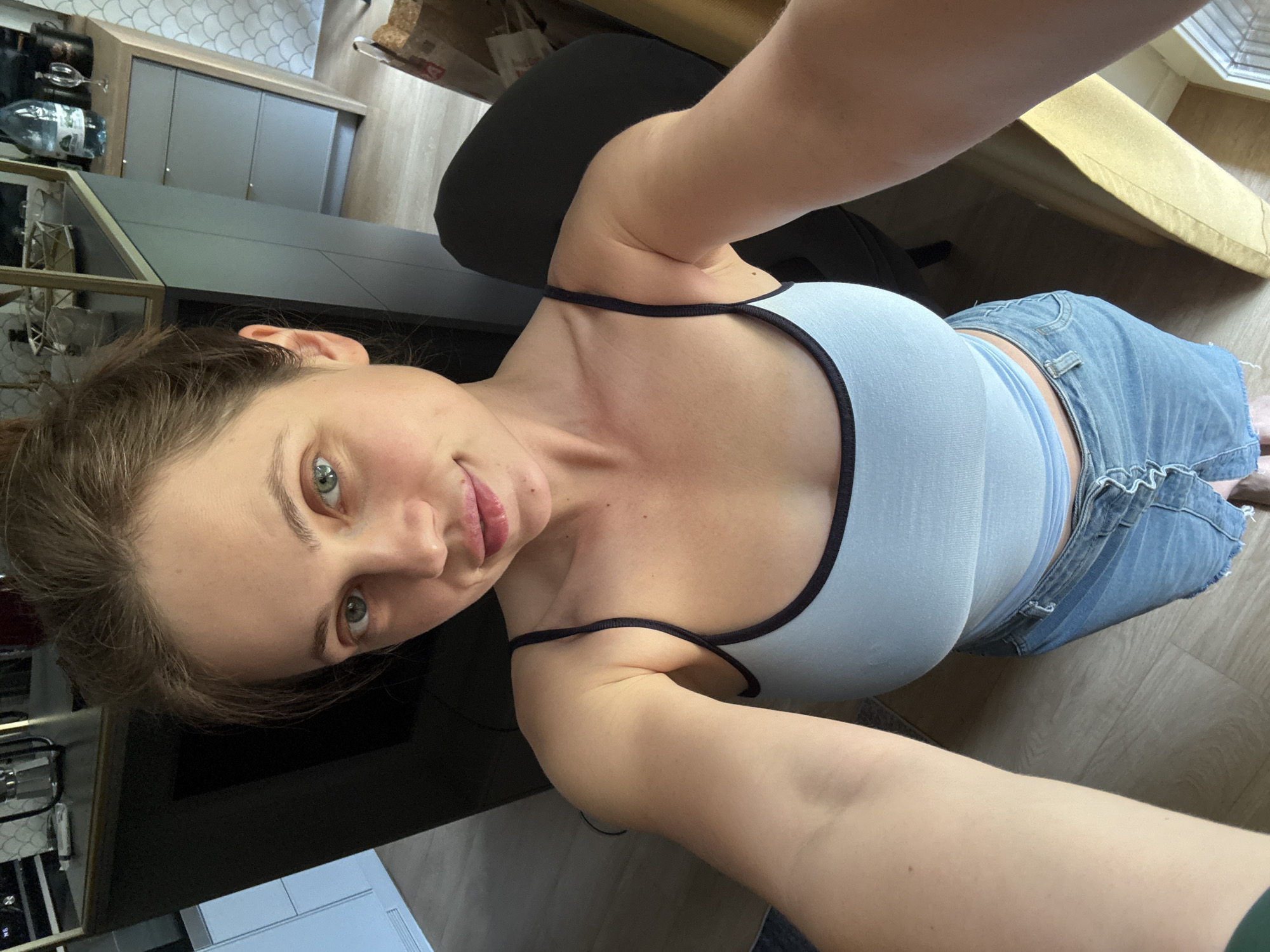The image shows a person taking a selfie in what appears to be a kitchen or dining area. The person, a woman with light skin and brown hair pulled back, is wearing a light blue tank top with black trim and denim shorts. She is looking towards the camera with a slight smile. The background includes kitchen cabinetry, a countertop, and a wooden floor. There is also a glimpse of furniture, possibly a table or chair, in the upper right corner. The lighting suggests it could be daytime. No visible text is present in the image.