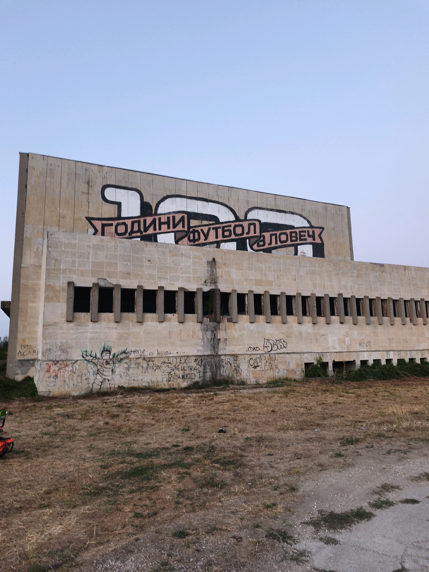 The image shows a large, brutalist concrete building in Lovech, Bulgaria, adorned with graffiti. The building has a long, multi-story facade with numerous rectangular openings that appear to be former windows or balconies, now empty and dark. A prominent banner-like graffiti painting stretches across the upper portion of the building, depicting stylized large numbers and text in Cyrillic. The text reads "100 ГОДИНИ ФУТБОЛ В ЛОВЕЧ," which translates to "100 Years of Football in Lovech."
The facade of the building is constructed from light-colored, roughly hewn stone blocks. Below the upper row of openings, the concrete wall is heavily covered with graffiti, including tag-like scribbles, a cartoonish figure, and more elaborate designs. Some of the graffiti is in Cyrillic and Latin scripts.
The ground in front of the building is covered with dry, overgrown grass and sparse vegetation. A dirt or gravel path runs in the foreground. Two red bicycles, one with a black seat and the other partially visible with orange accents, are parked on the left side of the image, near the edge of the building. The sky above is clear and a pale blue, indicating it is likely daytime. The overall impression is one of decay and abandonment, contrasted with the vibrant graffiti.