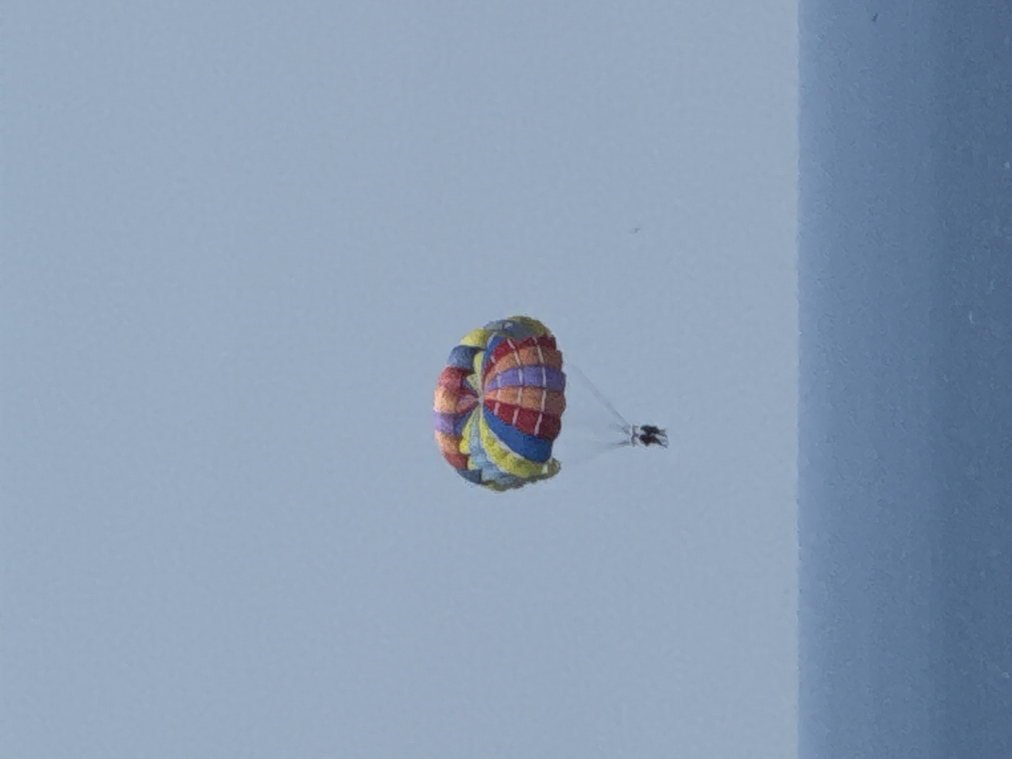 The image shows a person parasailing over the ocean. The parasail is multicolored and suspended in the sky against a pale blue backdrop. The ocean appears calm and blue. The scene is likely from a coastal area, possibly near Boreti, Montenegro, known for its beaches and water activities. There are no visible buildings or specific landmarks that would confirm the location in Boreti.
