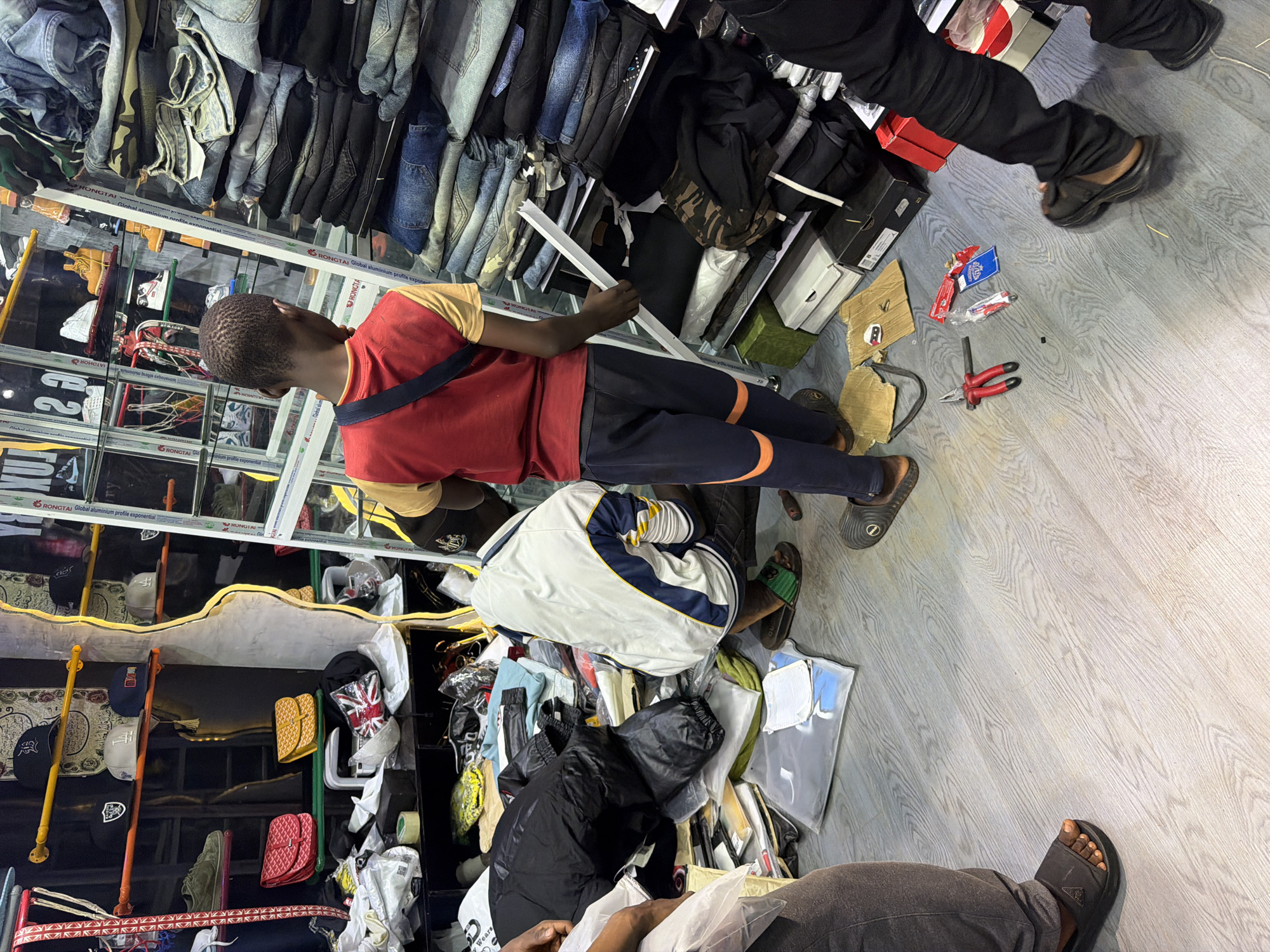 A boy in a red shirt and navy pants with orange stripes is standing in a store in Bukuru, Nigeria. He is looking down at a pile of clothes on the floor. To his left, another person in a white and blue jacket is kneeling on the floor, also looking at the clothes. The store appears to be a clothing store with shelves stocked with various items of clothing, including jeans, shirts, and jackets. There are also bags and shoes visible. A pair of pliers and some small items are on the floor near the boy. The floor is wooden.