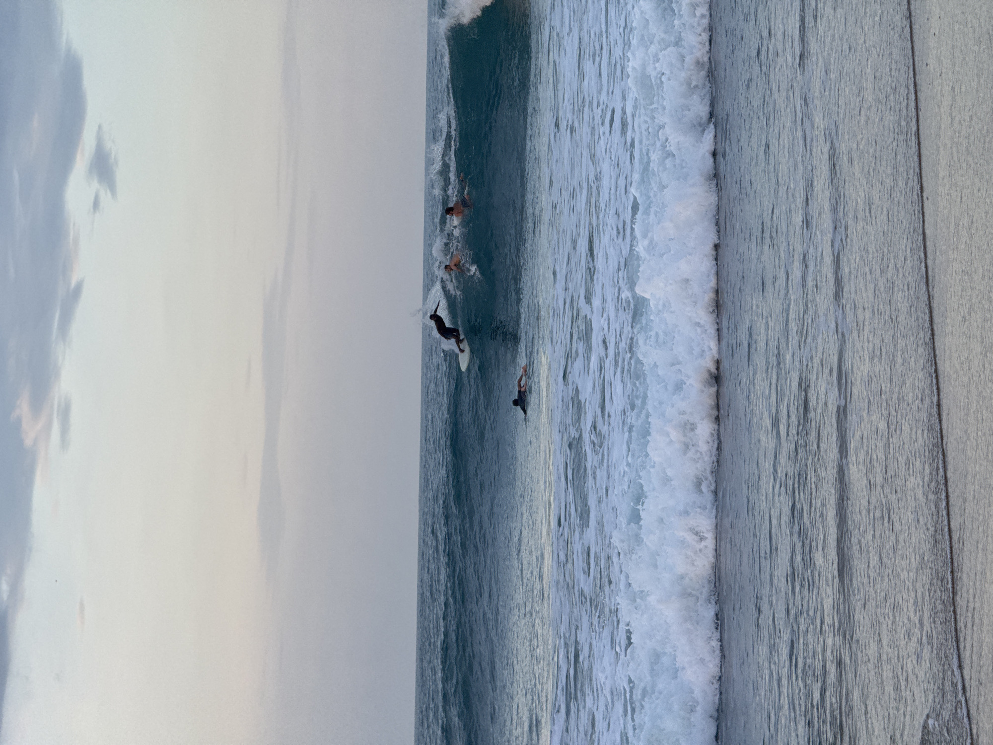 This image depicts a group of surfers in the ocean at Arugam Bay, Sri Lanka. The scene captures the activity of surfing during what appears to be late afternoon or early evening, given the soft, diffused light and the muted tones of the sky. The weather seems calm, with a gently overcast sky that doesn't obscure the view of the ocean.
Several surfers are visible in the water. One surfer in the foreground is actively riding a wave on a white surfboard, leaning into a turn. Further back, other surfers can be seen paddling or waiting for waves, some partially submerged in the water. The ocean itself is a deep blue-green, with white foam cresting on the breaking waves. The texture of the water is prominent, showing the movement and power of the waves.
To the right of the frame, a portion of the shoreline or a man-made structure, possibly a seawall or pier, is visible. This structure has a textured surface and a lighter, grayish-blue color, contrasting with the dark water and the softer tones of the sky. The overall impression is one of a serene yet active coastal scene, highlighting the sport of surfing in a beautiful natural setting. There is no visible text in the image.