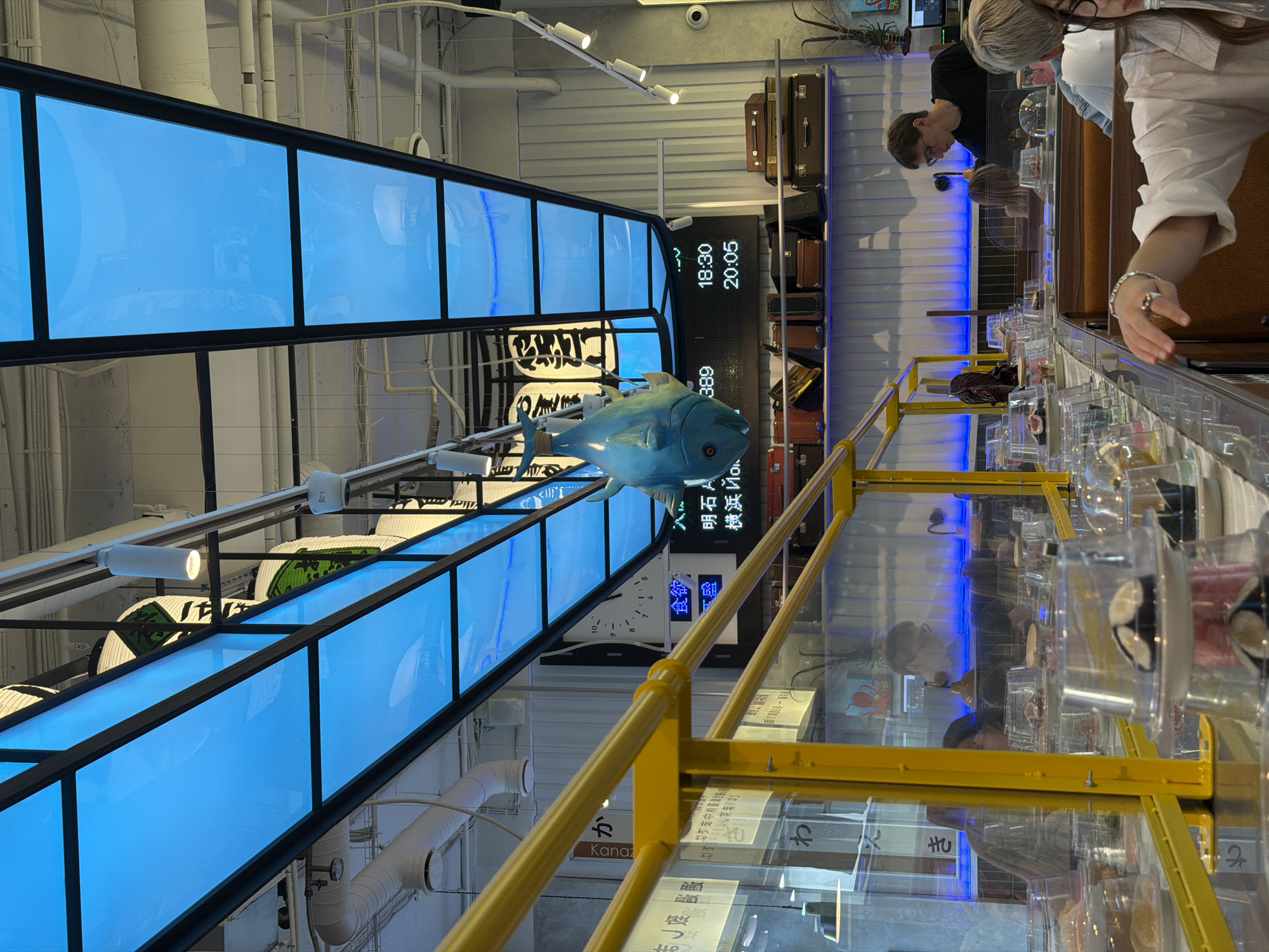 A conveyor belt sushi restaurant is depicted, with a blue illuminated ceiling and a decorative blue fish hanging from the ceiling. Several plates of sushi are visible on the conveyor belt, protected by clear plastic lids. People are seated at the restaurant, some of whom are visible through transparent partitions. The setting appears to be a modern, interior space with yellow accents. The location in Saint Petersburg, Russia, is not evident from the visual information provided in the image.