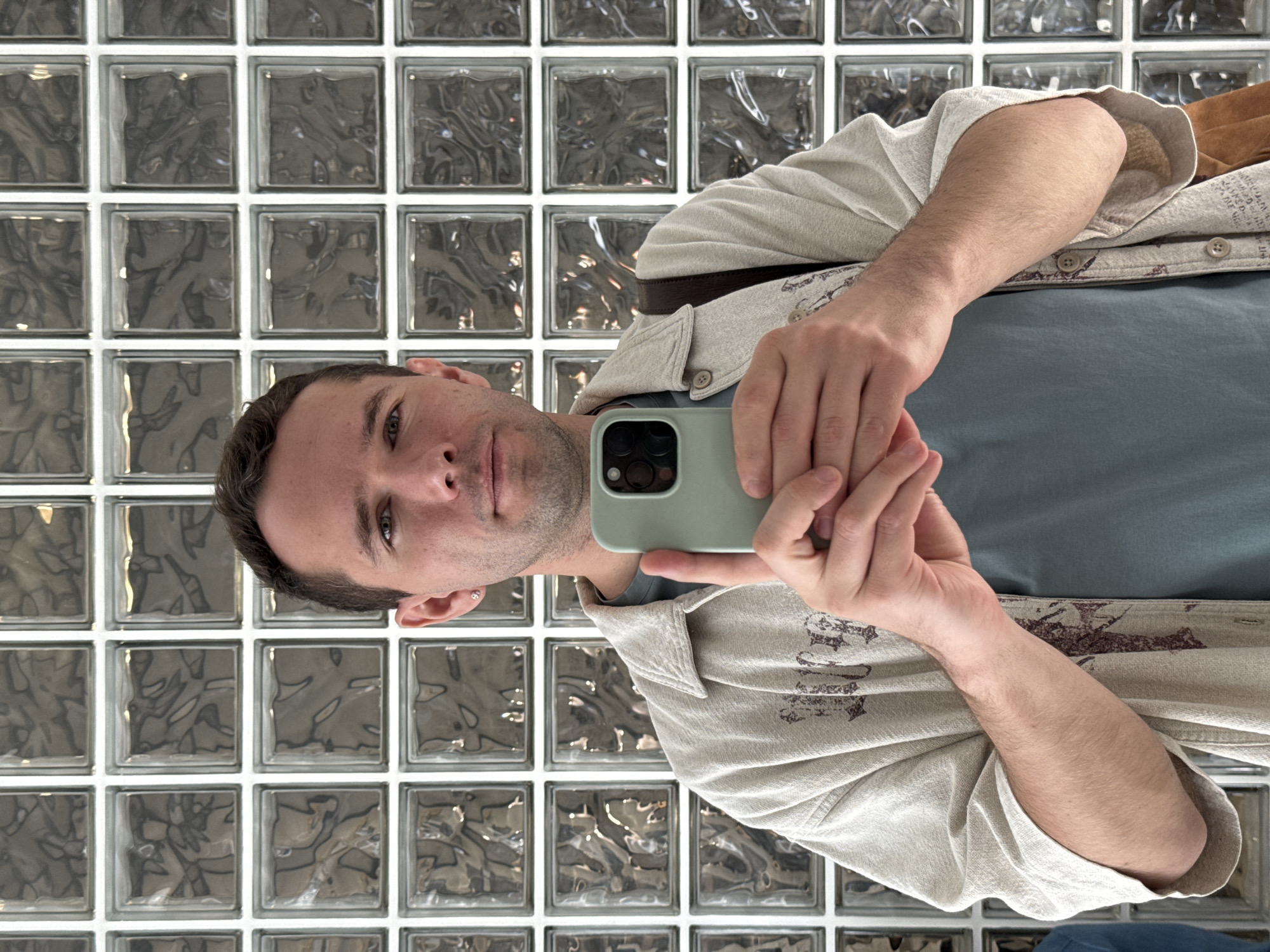 A man is taking a selfie in front of a glass brick wall. The man is wearing a light grey button-down shirt over a blue t-shirt and has a brown belt visible. He is holding a light green smartphone. The glass brick wall is made of clear glass bricks with a textured pattern. The location is Moscow, Russia.