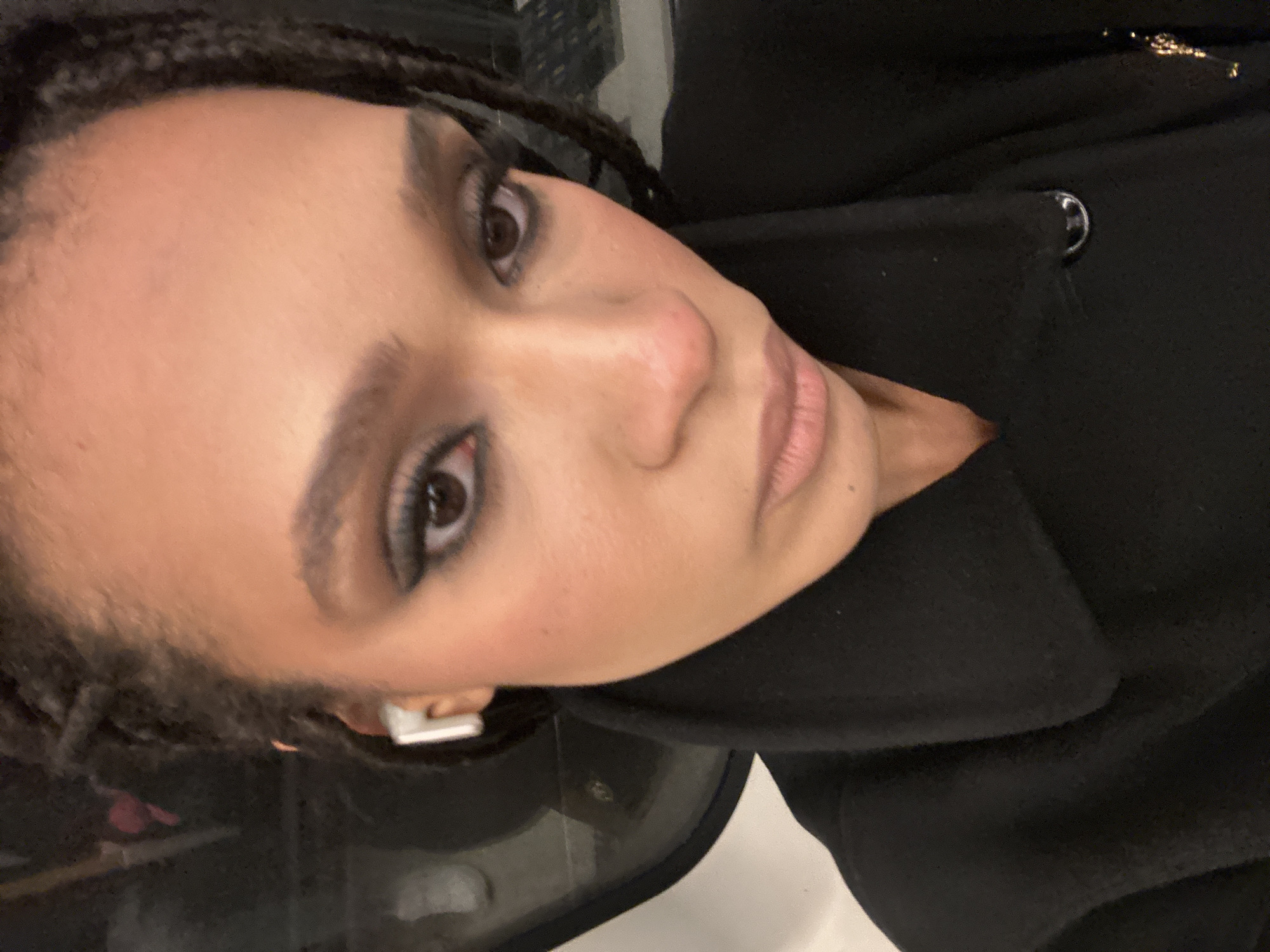 A close-up, slightly angled portrait shows a person with brown eyes and braided hair, wearing dark eye makeup, and wearing an earbud in their left ear. They are wearing a black coat, which covers most of the frame. There's a metallic button on the coat's lapel. The setting appears to be indoors, possibly on public transport, given the reflective surface and potential window visible to the left. The location cannot be determined to be in London.