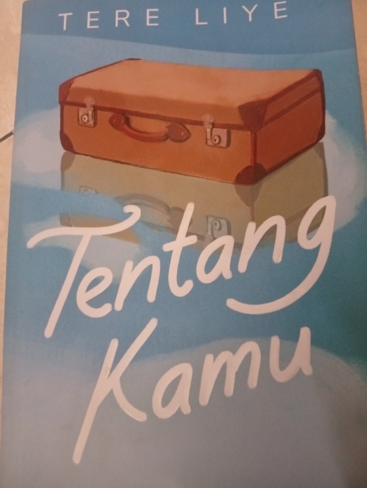 The image is a close-up of the cover of a book titled "TERE LIYE" with the words "Tentang Kamu" written in a large, flowing script below. The background is a light blue gradient, suggesting a sky or clouds. On the upper portion of the cover, a stylized illustration of a brown suitcase with red trim and a red handle is depicted. Below the suitcase, a faint reflection or silhouette of another, lighter colored suitcase is visible. The text and illustration are rendered in a way that suggests a painterly or illustrative style. There are no people or any identifiable landmarks or settings in the city of Bajingjowo, Indonesia, within this image.