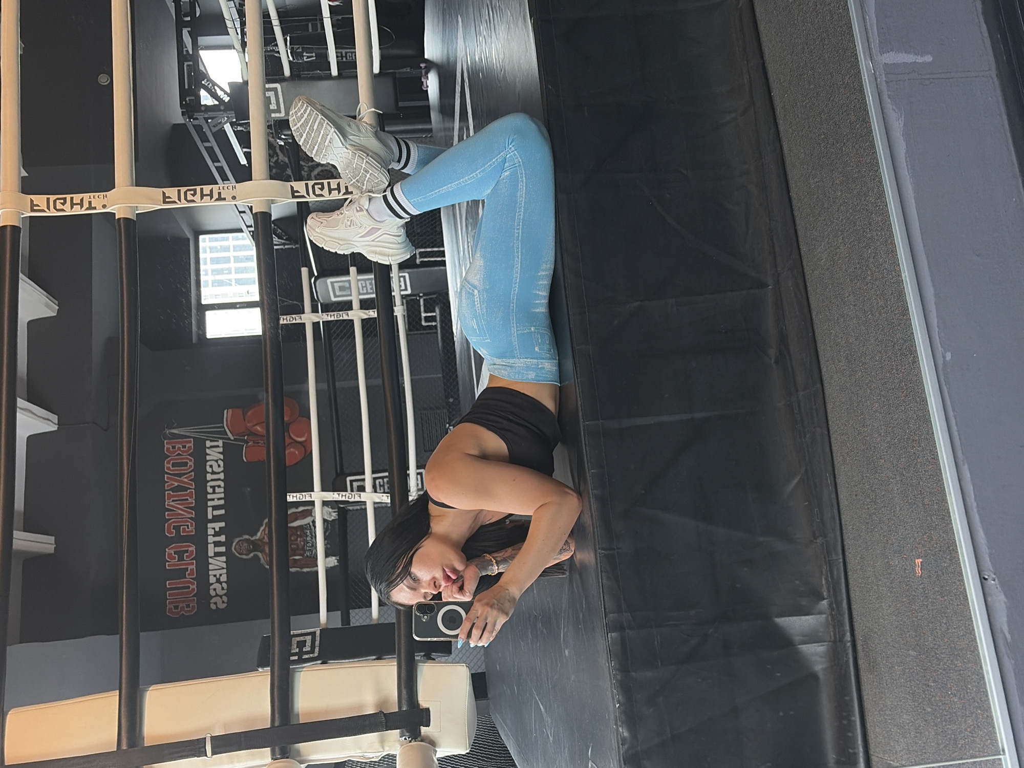 The image shows a woman in a boxing ring, taking a selfie in an upside-down position. She is wearing light blue leggings with a denim-like pattern, a black tank top, white athletic shoes, and white socks with black stripes. The woman has long dark hair, visible tattoos on her arm, and is wearing jewelry. She is holding a smartphone to take the photo.
The setting appears to be an indoor boxing gym. The background shows the ropes of a boxing ring, with white and dark vertical posts. A sign on the wall reads "BOXING CLUB" in red letters. The floor of the ring is covered with black material. The lighting suggests it is daytime, possibly with artificial lighting supplementing natural light from windows visible in the background. The woman's expression is neutral, and the overall mood is focused on fitness and training. The provided location context is Ufa, Russia.