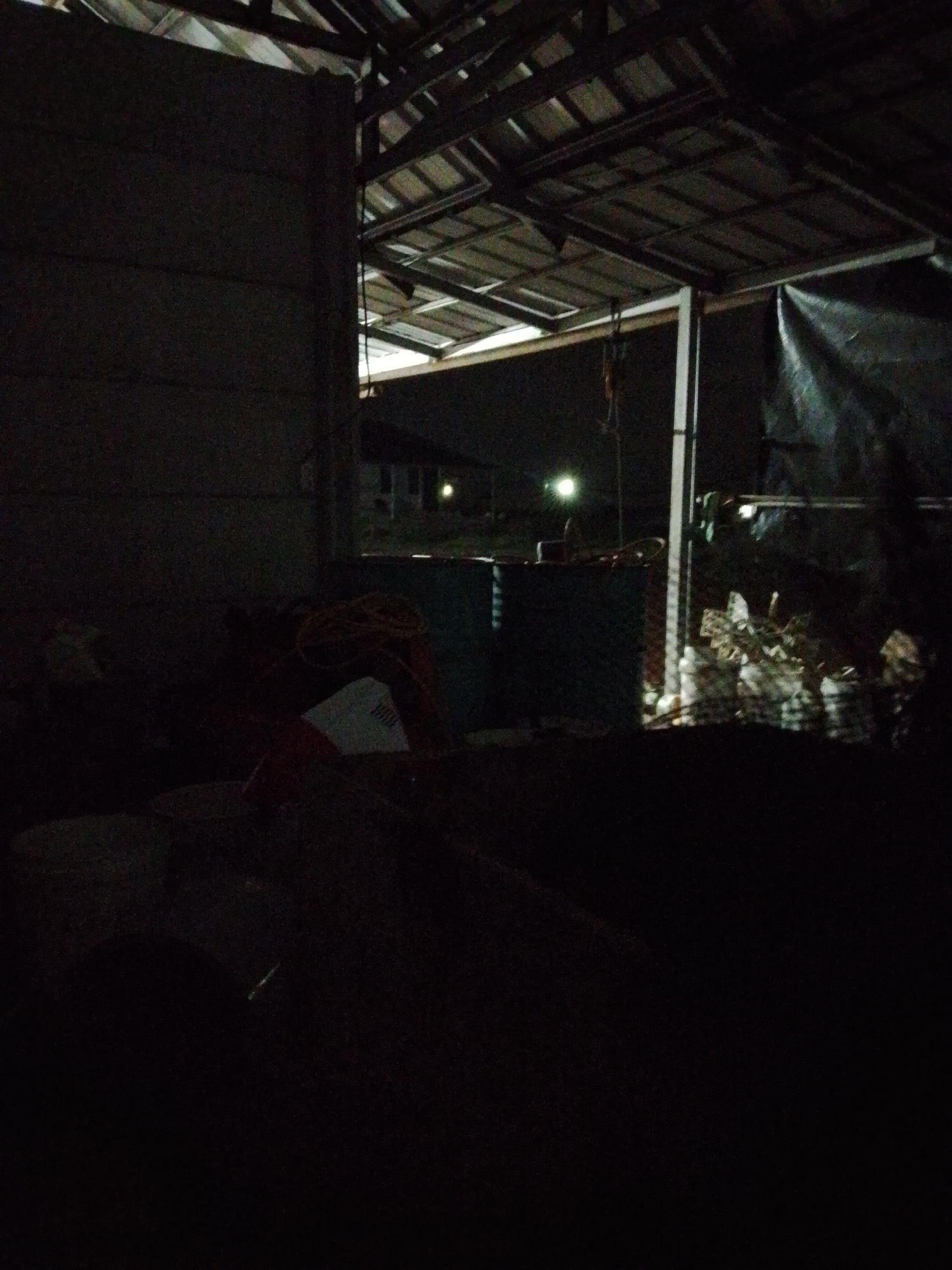 The image appears to be taken at night in what looks like an open barn or shelter structure in Bukit Ketok, Indonesia. The barn has a metal roof supported by beams and white metal poles. The interior is mostly obscured by darkness. In the distance, there are lights and the outline of a building, possibly a house. Inside the barn, various objects are barely visible, including what appears to be buckets or containers, and a dark tarp. The foreground is completely black, with no discernable details.